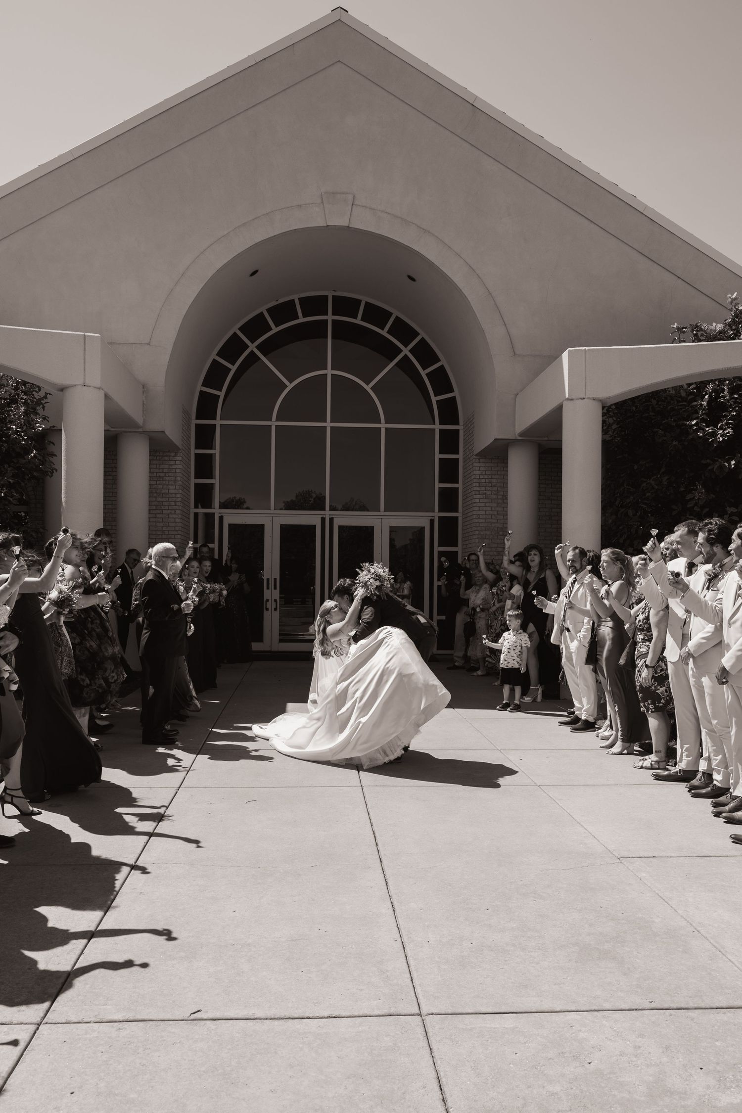 Black and white photo of bride and groom sharing a dip kiss outside a church surrounded by cheering wedding guests.
