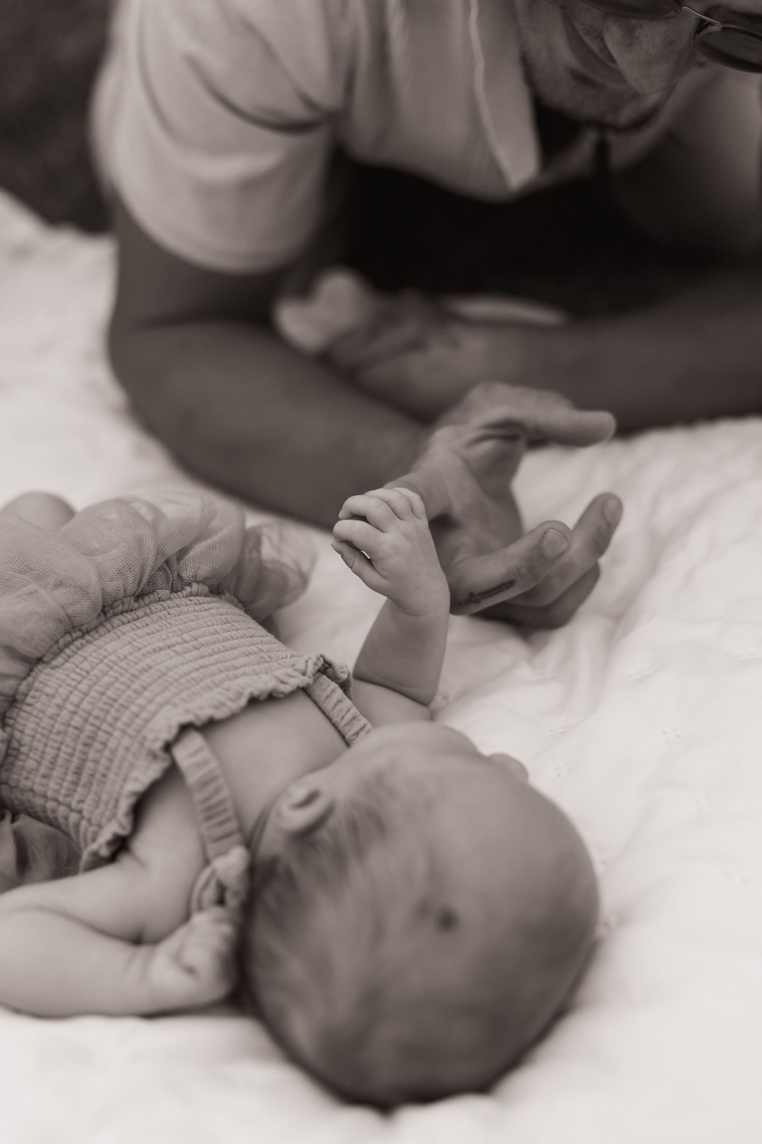 Black and white photo of a newborn baby lying on a white blanket while an adult gently holds its tiny hands.