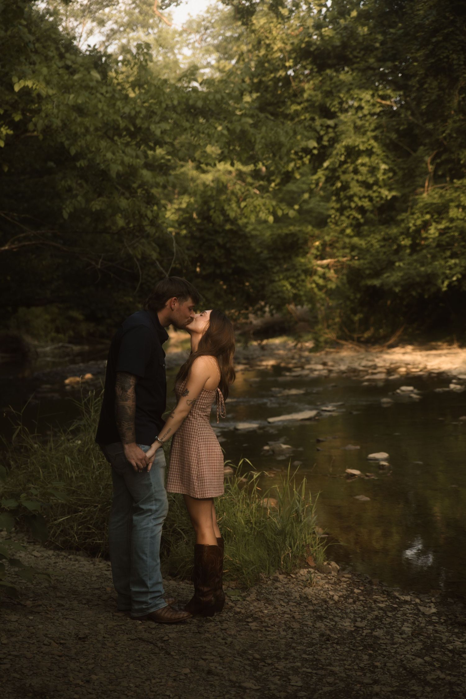 Couple sharing a romantic kiss by a serene creek surrounded by lush green trees in warm golden light.