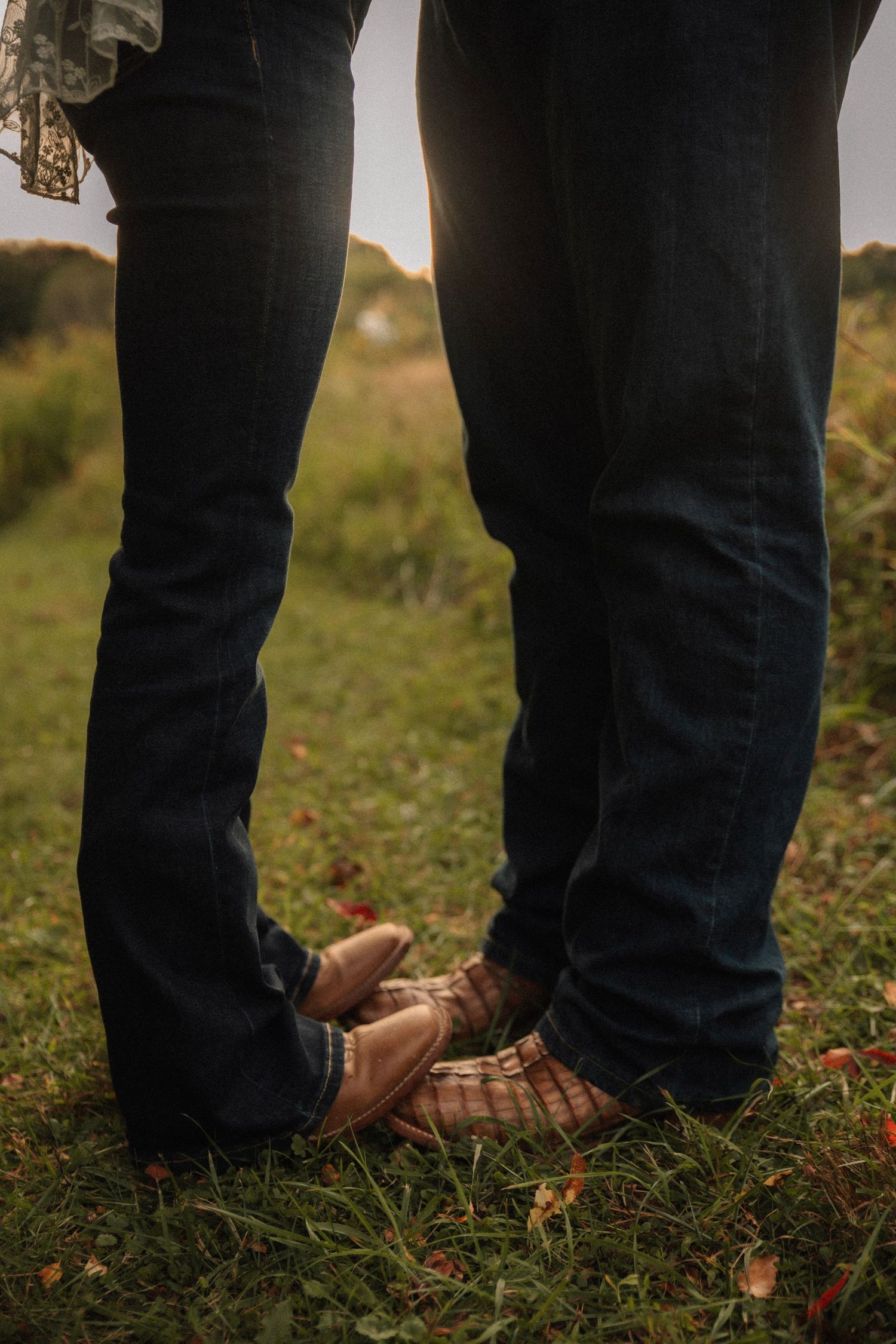 Two pairs of feet stand close together on grass, one wearing brown leather shoes and dark jeans.