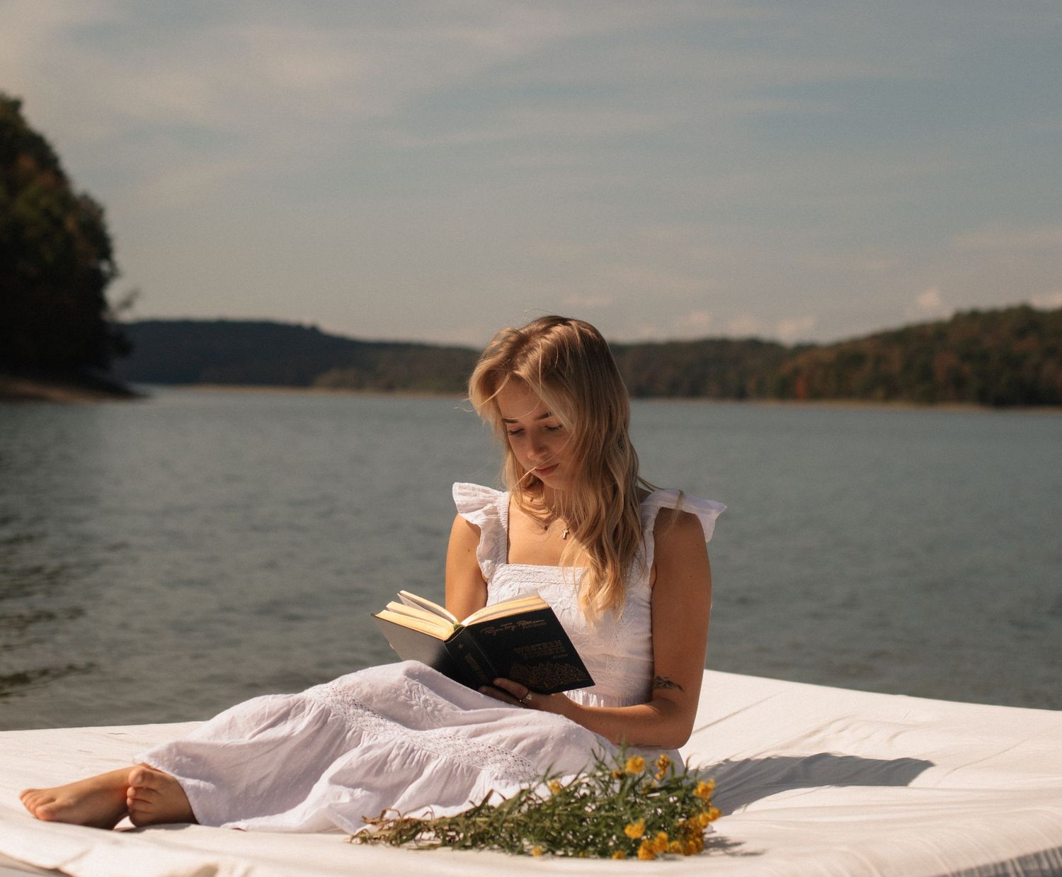A person in a white dress reads a book while relaxing on a blanket by a serene lake on a sunny afternoon.