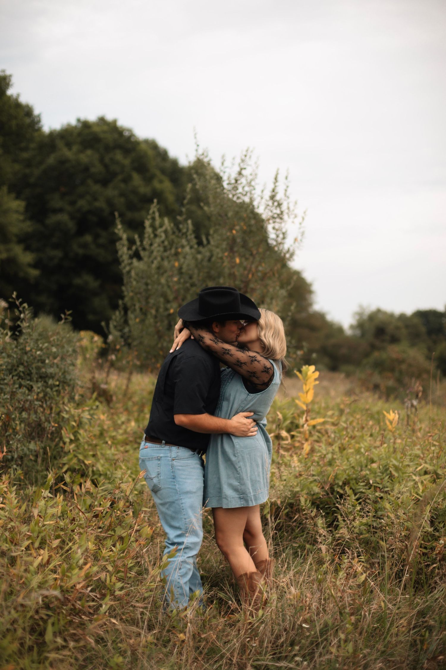 Couple kissing in a wildflower field, man wearing black cowboy hat and jeans, woman in blue dress, surrounded by lush greenery.