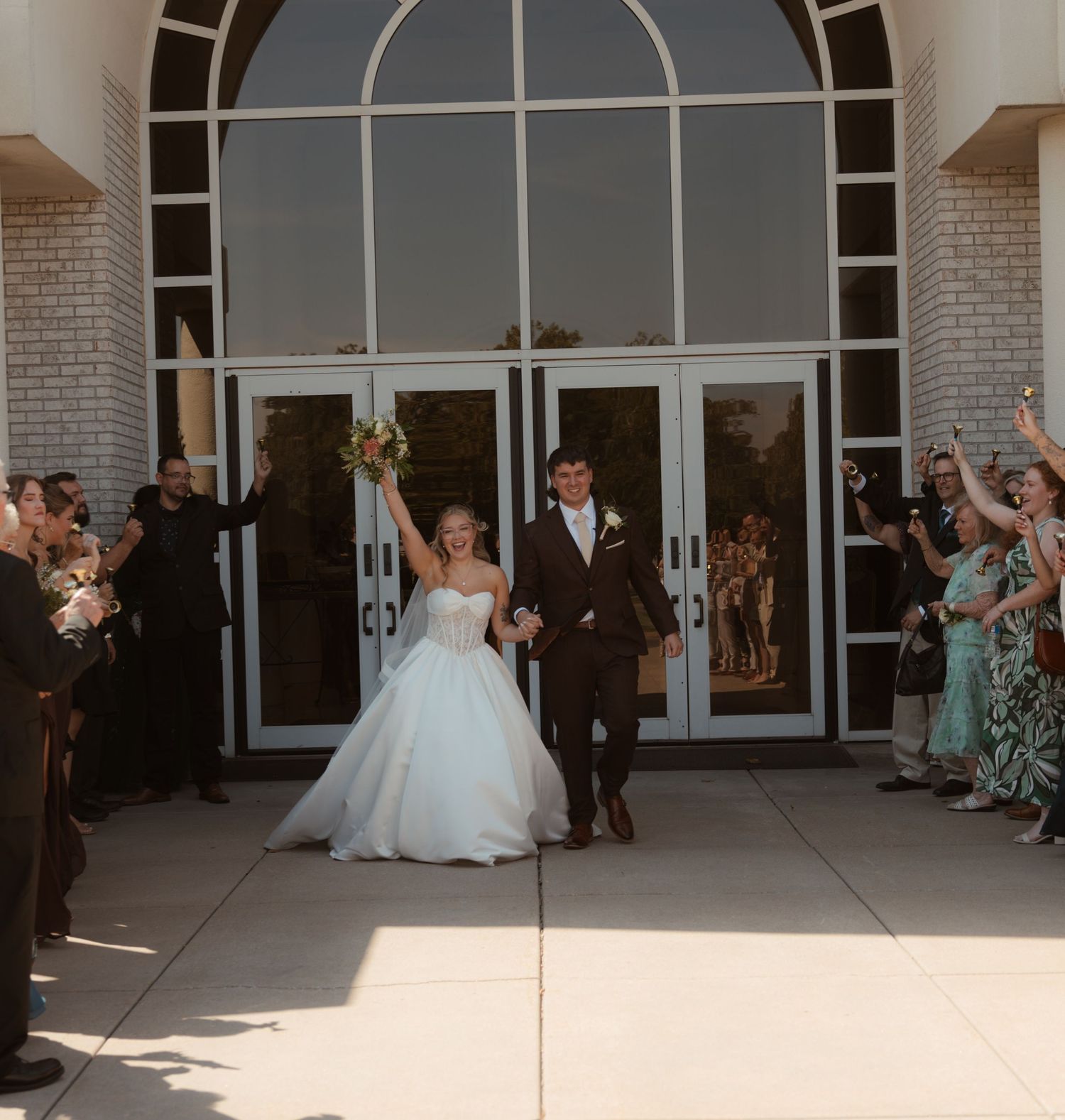 Bride and groom exit church doors celebrating with raised hands as wedding guests cheer along the walkway.
