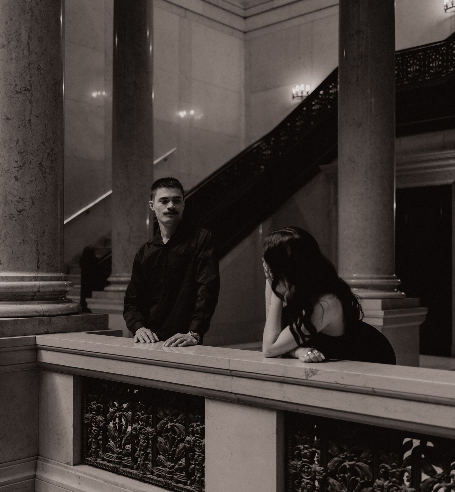 A man and woman in black attire lean on an ornate railing in a grand classical building with columns.