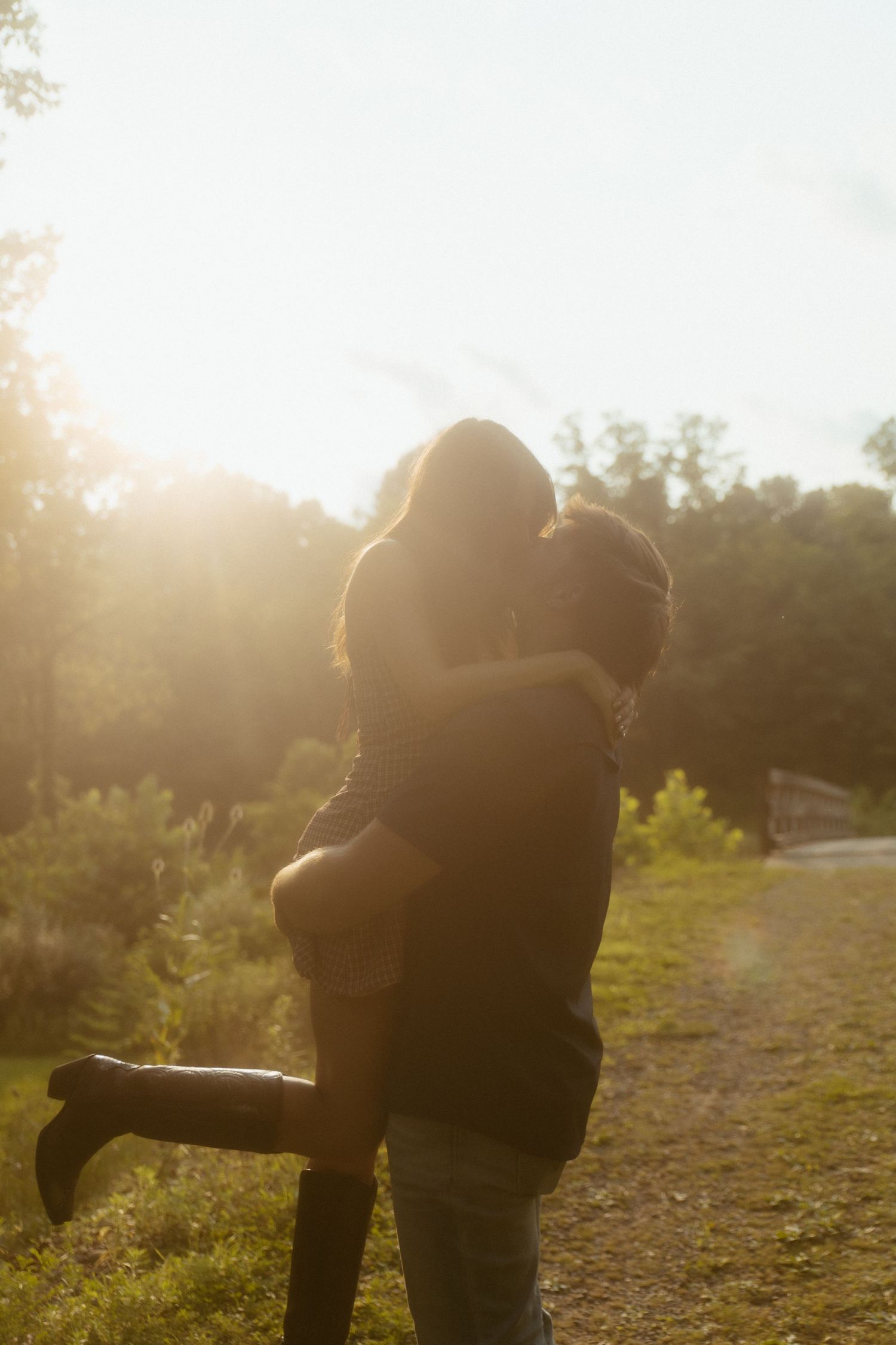 Couple sharing a romantic kiss in golden sunset light on a scenic park path surrounded by lush green trees.