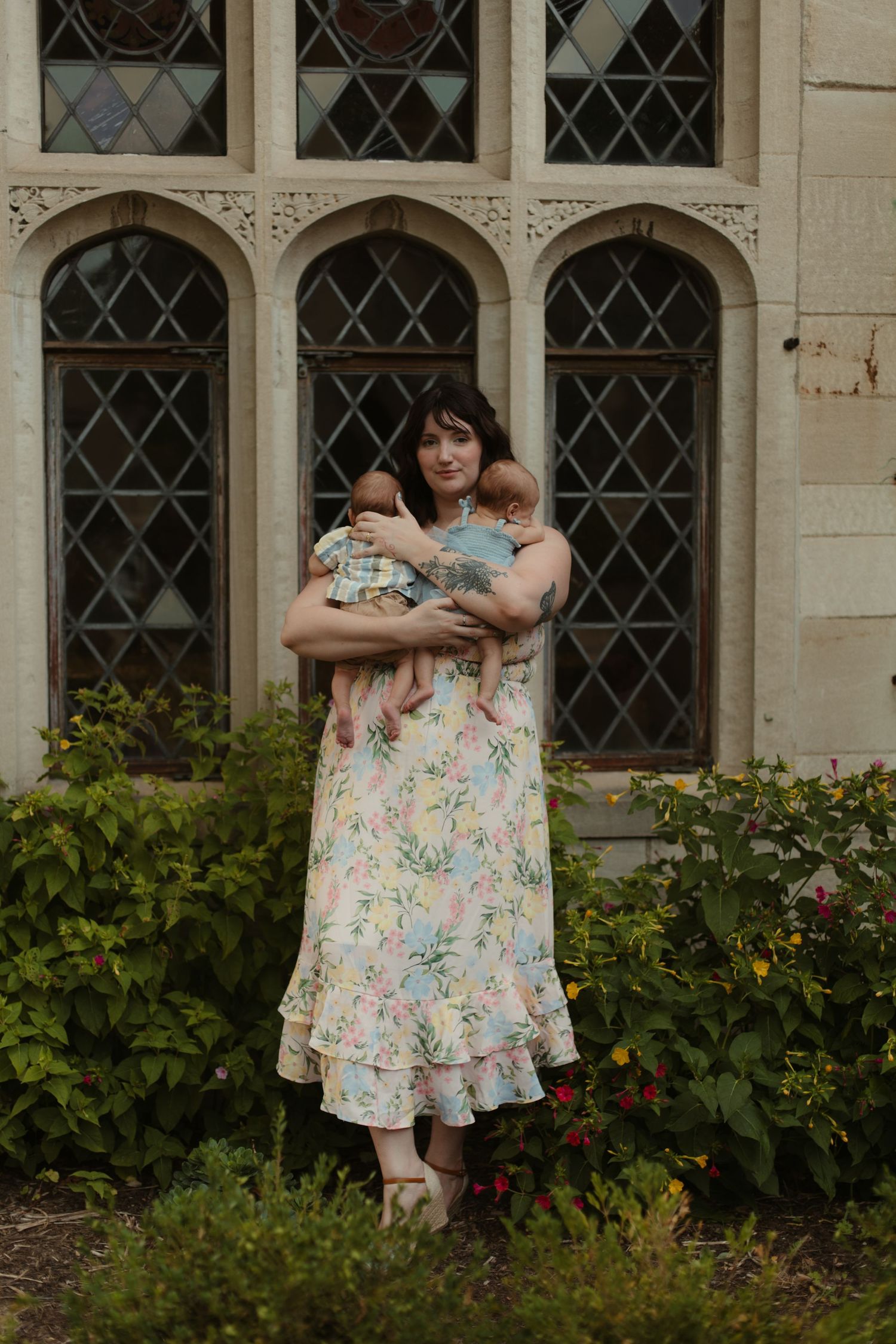 Woman in floral dress holding two babies outdoors near arched windows and lush greenery.