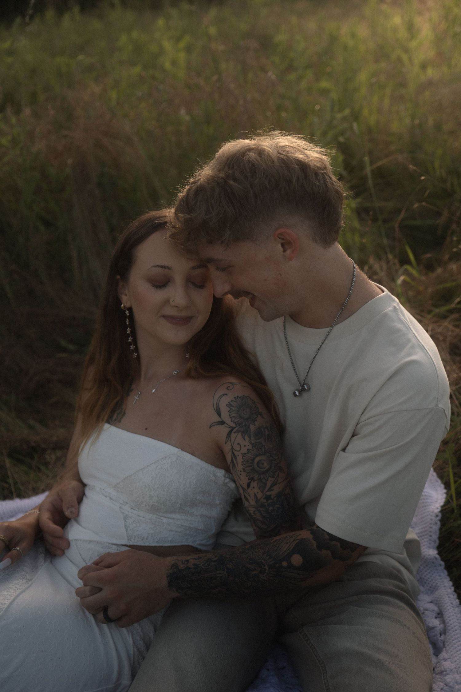 Couple sitting in a sunlit meadow, man with tattoos embracing woman in white dress at golden hour.