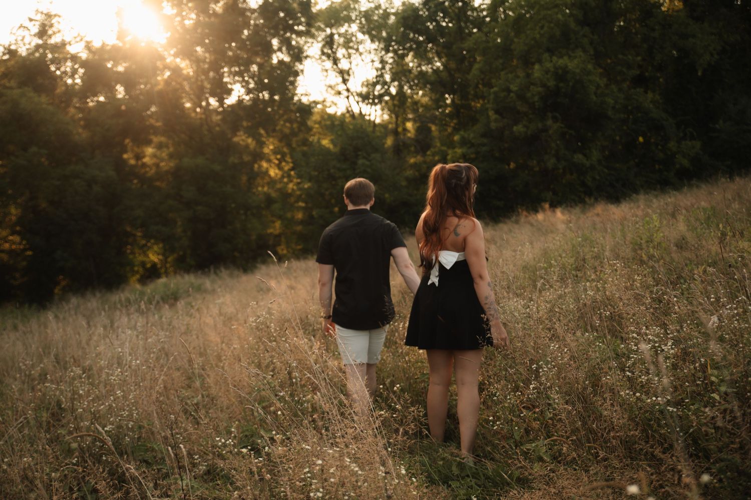 Couple walking through a golden sunlit meadow at sunset holding hands in a romantic outdoor setting.