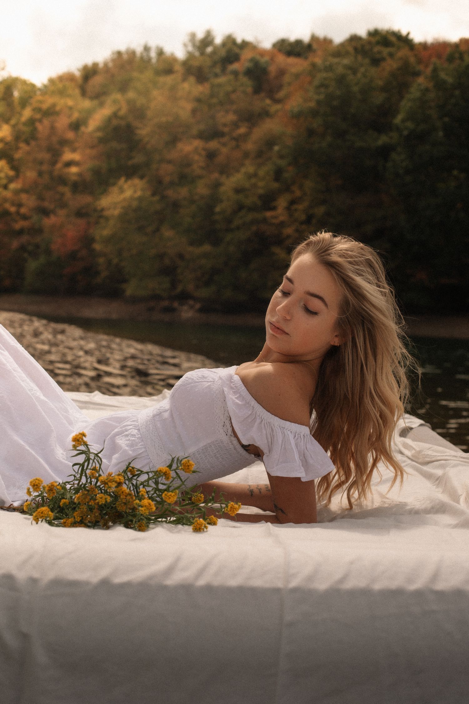 Woman in white dress lying on blanket by autumn lake with sunflowers at sunset.