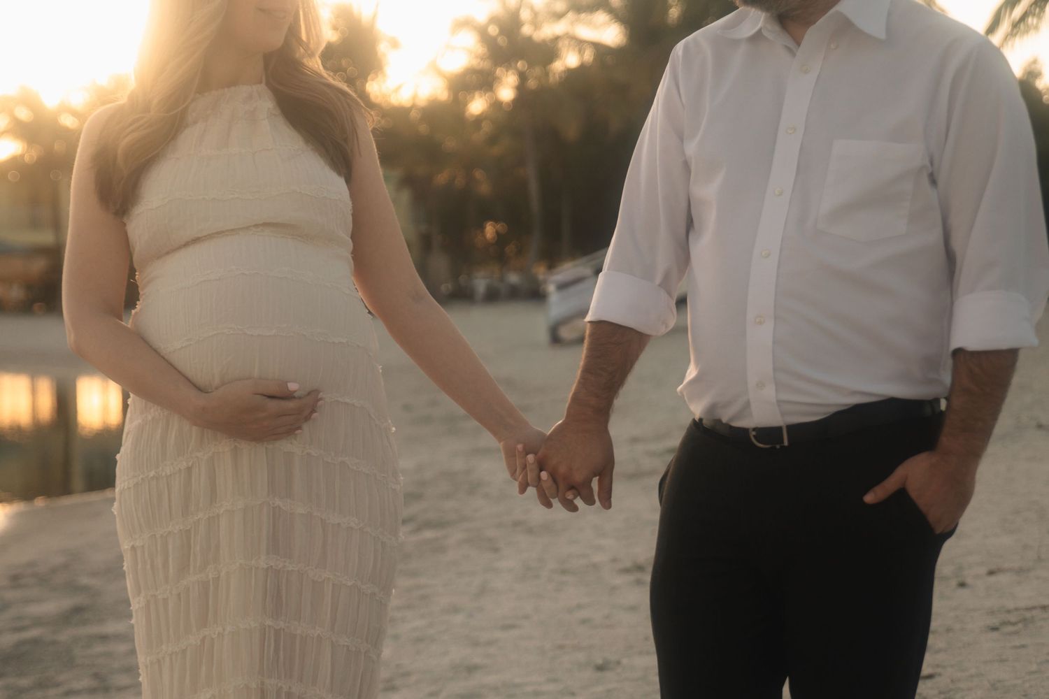 A couple in white outfits poses together on a sandy beach at sunset during a romantic photoshoot.