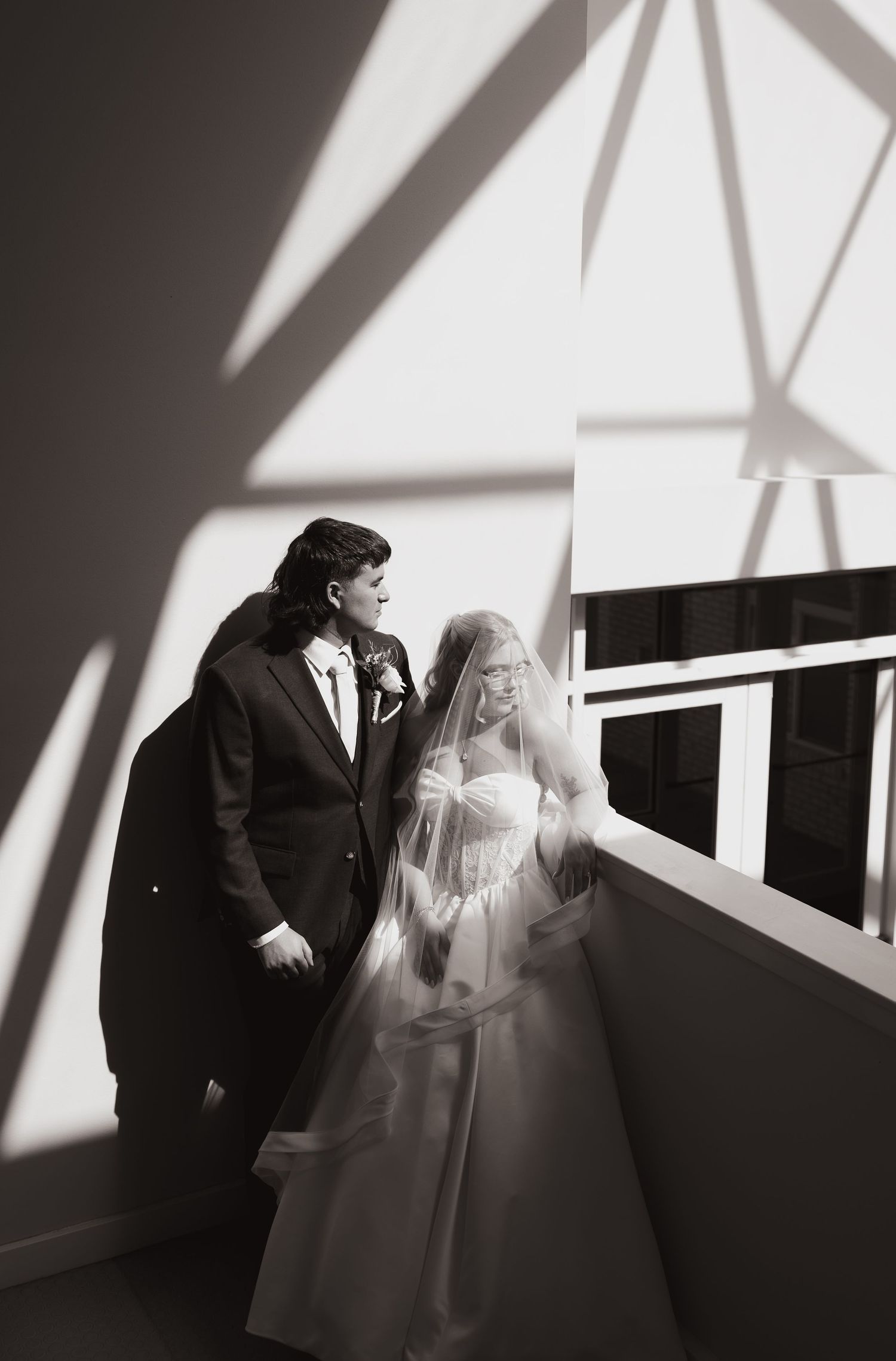 Black and white wedding photo of bride in veil and groom in suit standing by sunlit window.