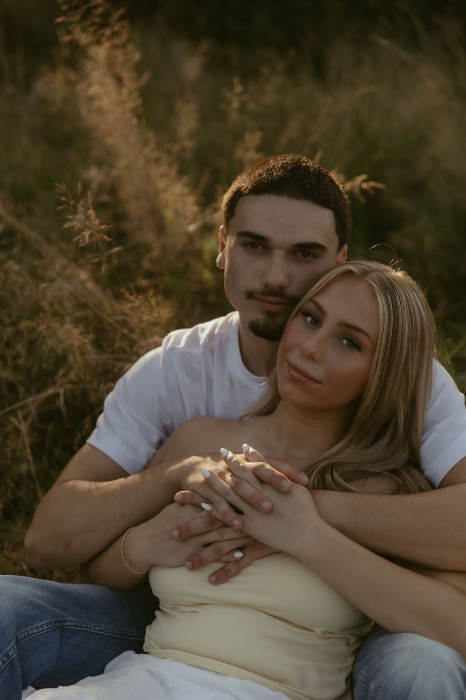 A couple sits together in a golden field, the man embracing the woman from behind as they hold hands romantically.
