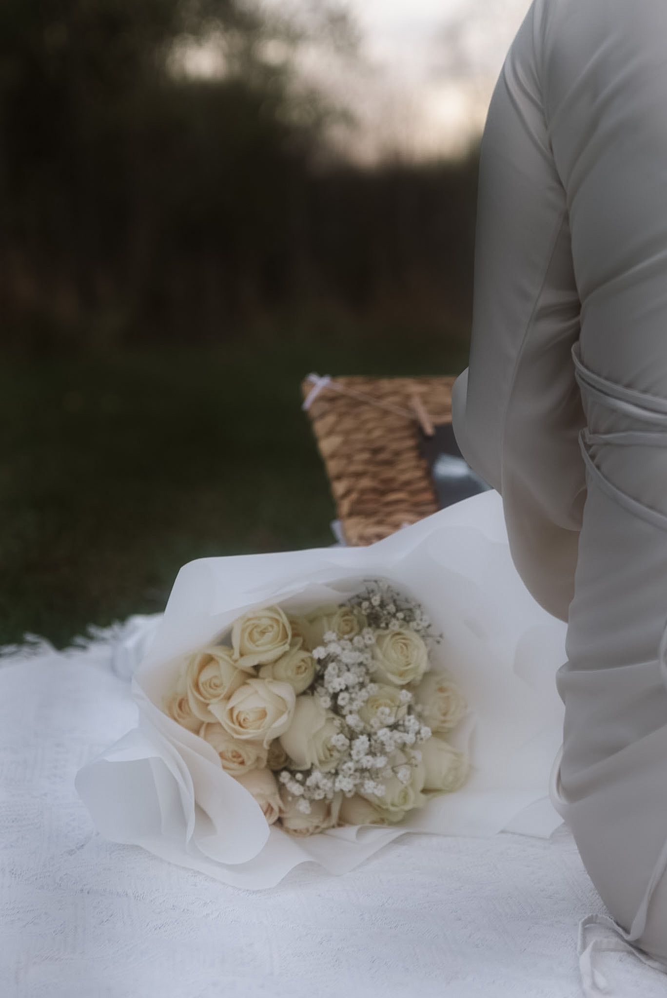 Person in gray dress holding white rose bouquet wrapped in white paper on a blanket outdoors.