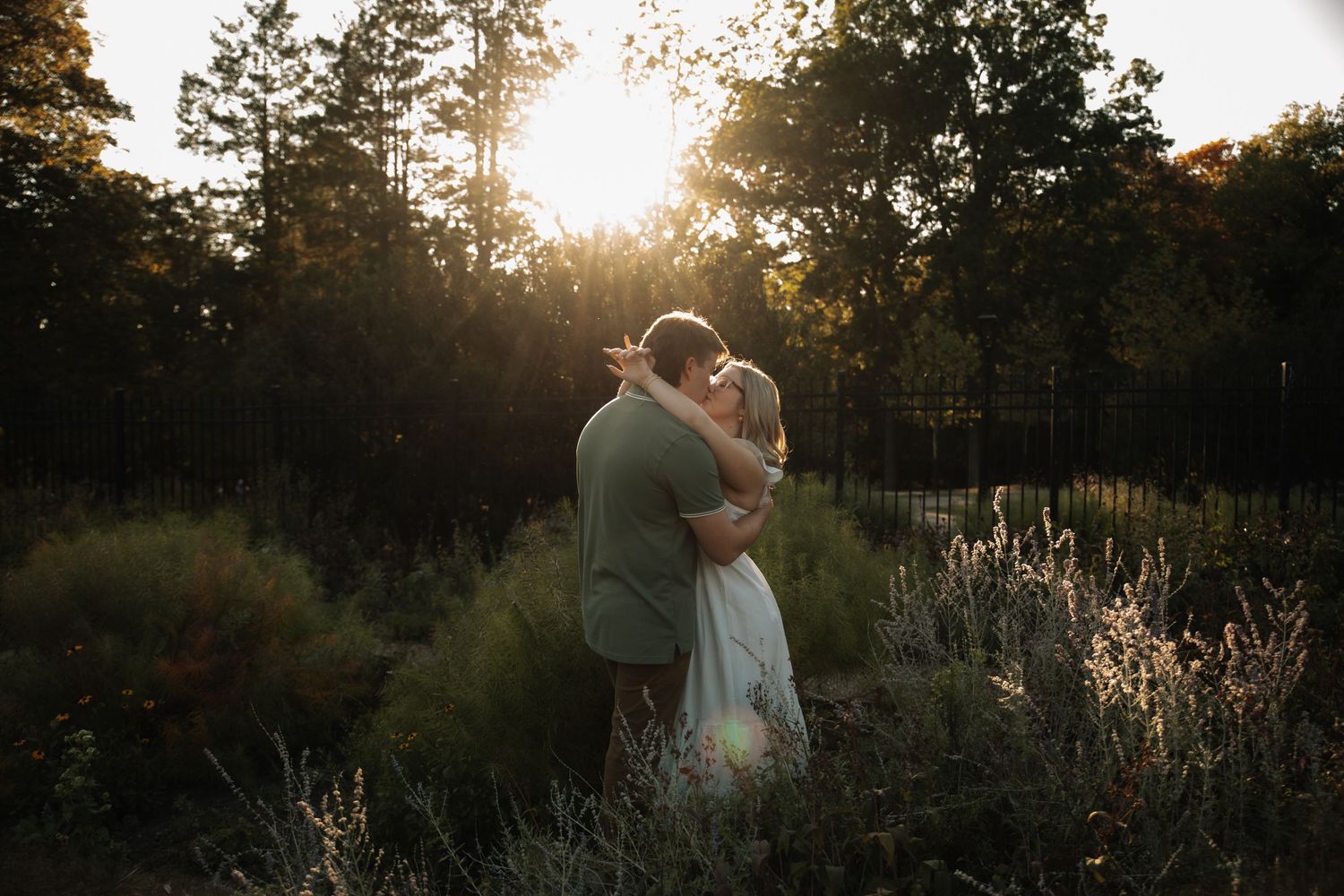 A couple embraces in a golden sunset among tall grass and trees in a romantic outdoor setting.