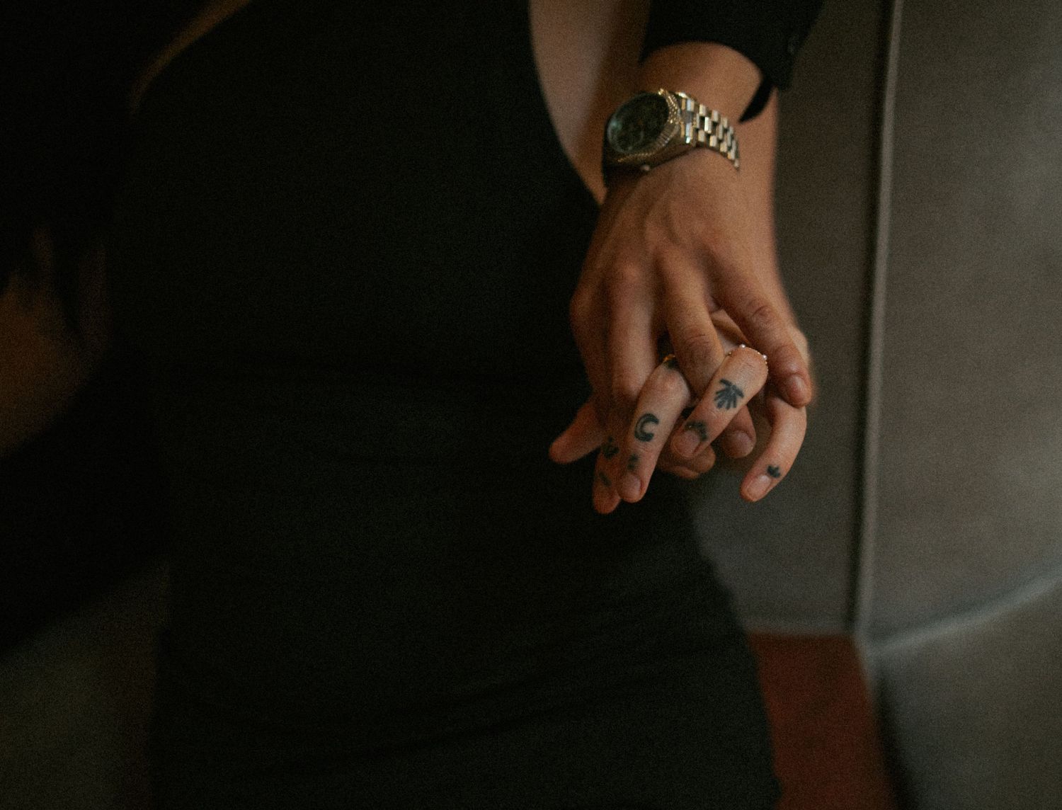 Close-up of hands with finger tattoos and a gold watch, clasped together against a black dress.