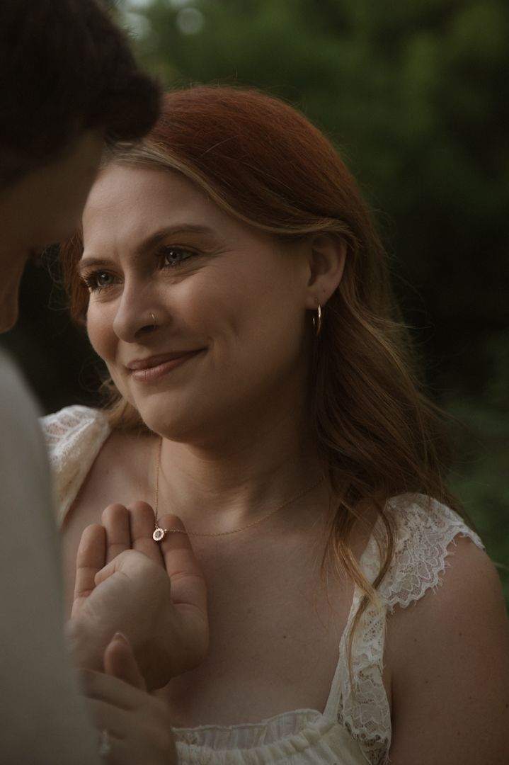 A smiling redhead woman in a white lace dress holds hands with someone while wearing a delicate necklace outdoors.