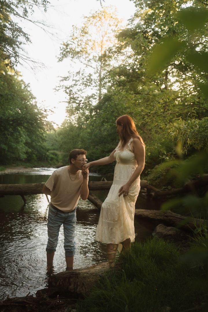 A couple shares a romantic moment by a sunlit forest river, man kissing woman's hand as she stands on a log.