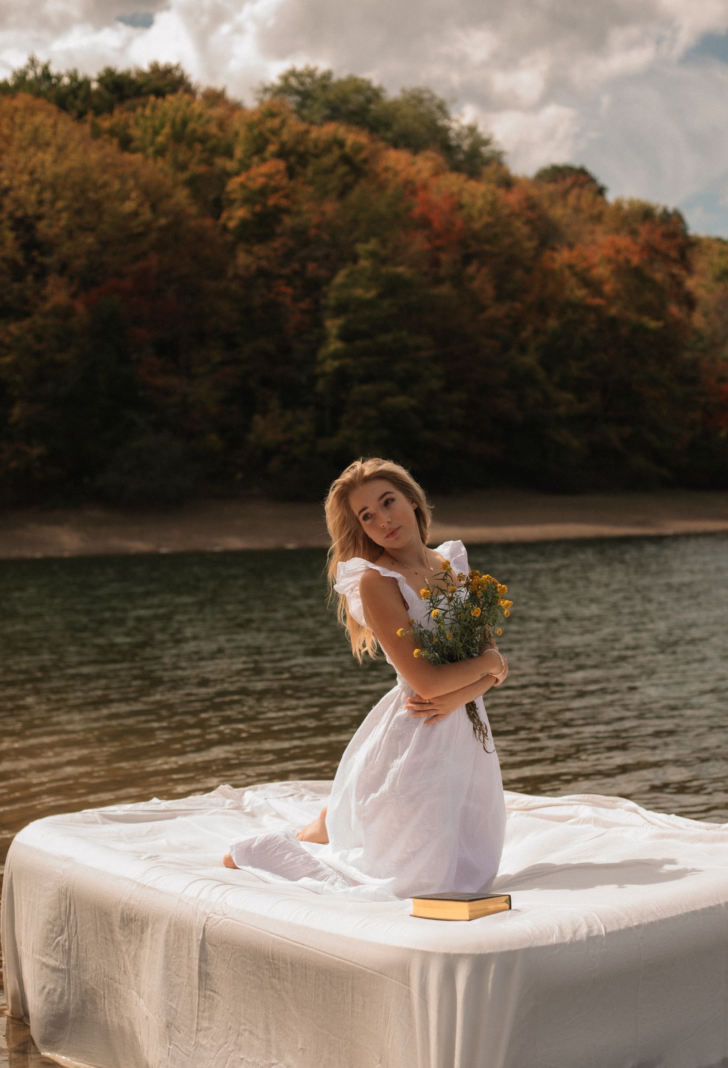 Woman in white dress holding wildflowers sits by autumn lake with colorful foliage backdrop.