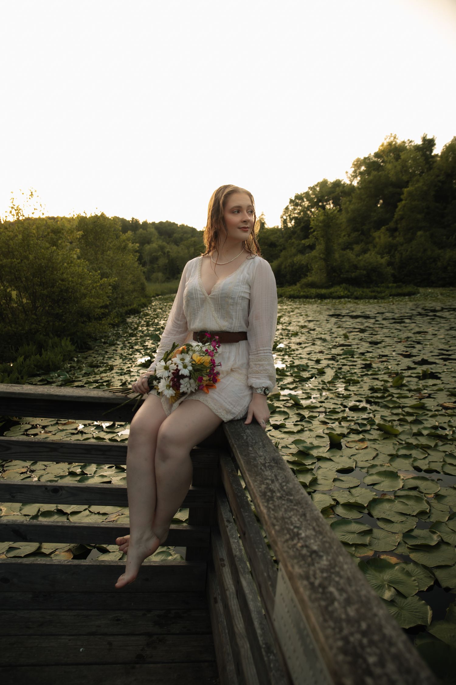Young woman in white dress holds wildflowers while sitting on wooden bridge over lily pad pond at sunset.