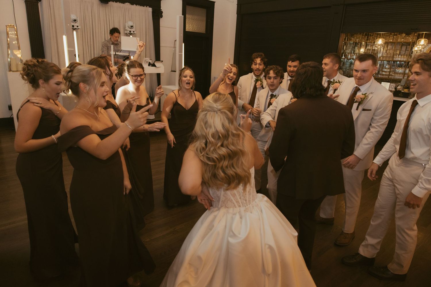 Bride in white gown surrounded by bridesmaids in black dresses and groomsmen celebrating on wedding reception dance floor.