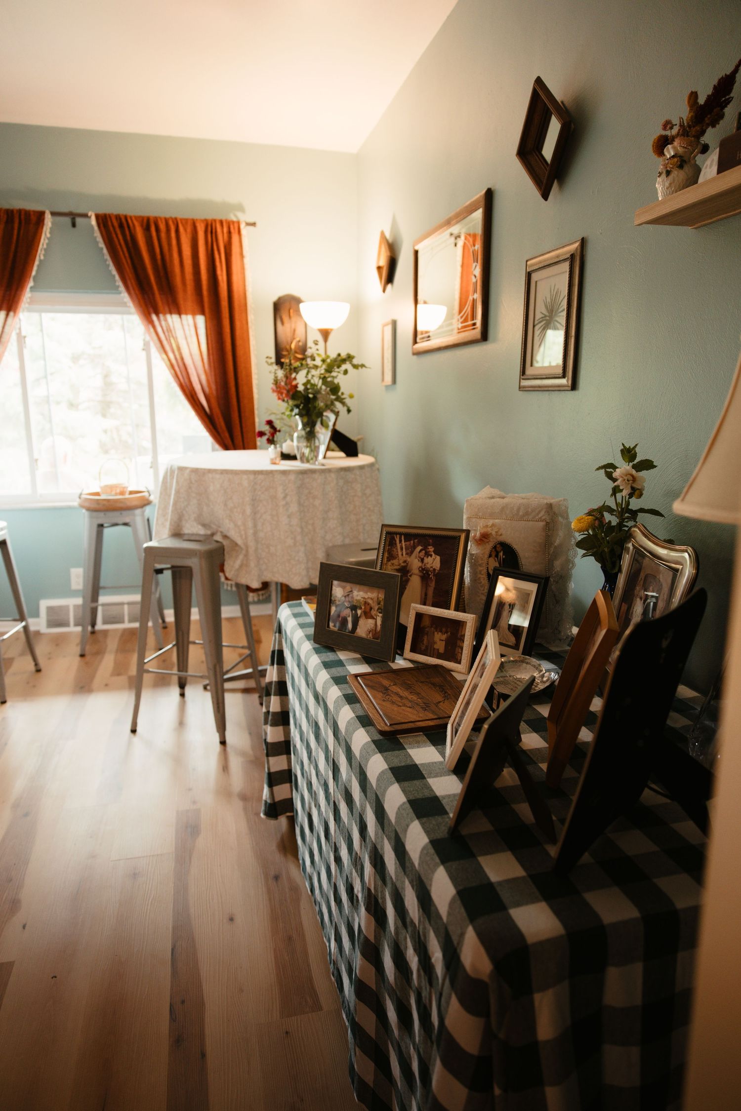 Cozy dining room with plaid tablecloth, family photos, rust curtains, and sage green walls with wood floors.