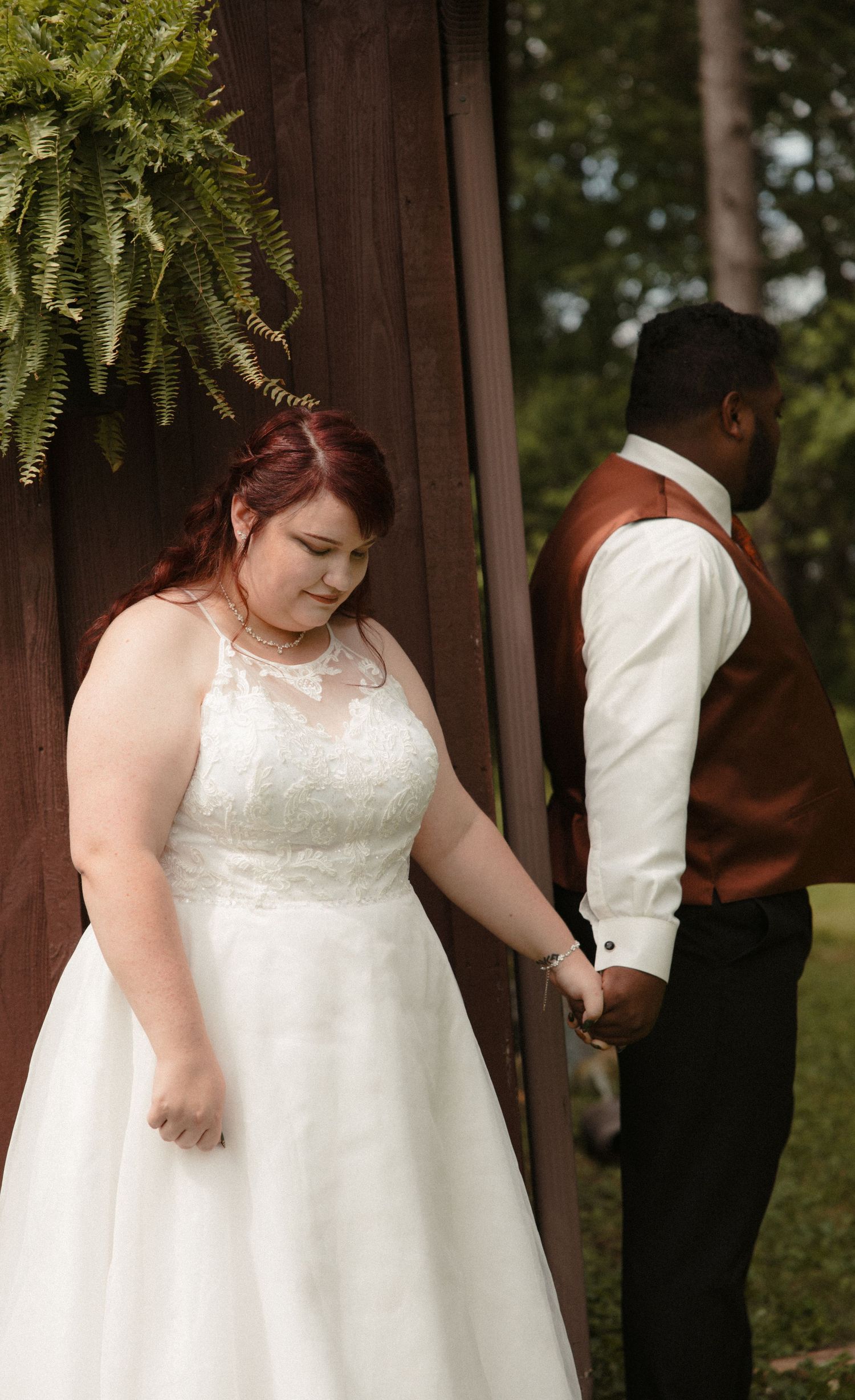 A bride in a white lace gown holds hands with a groom in a brown vest during an outdoor wedding ceremony.