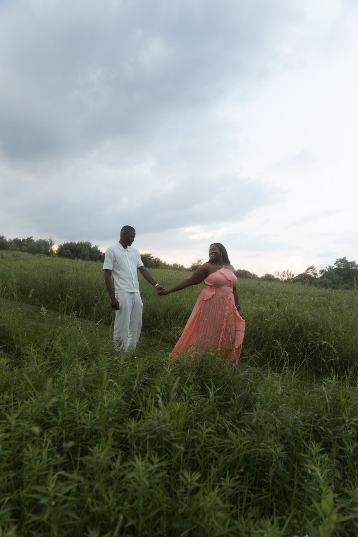 A couple holds hands in a lush green field, woman in pink dress, man in white, under a cloudy sky.