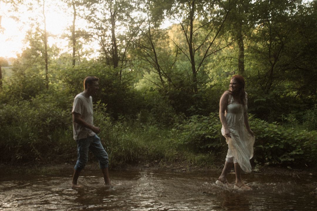 Couple wading through a shallow creek surrounded by lush green trees in warm golden sunset light.