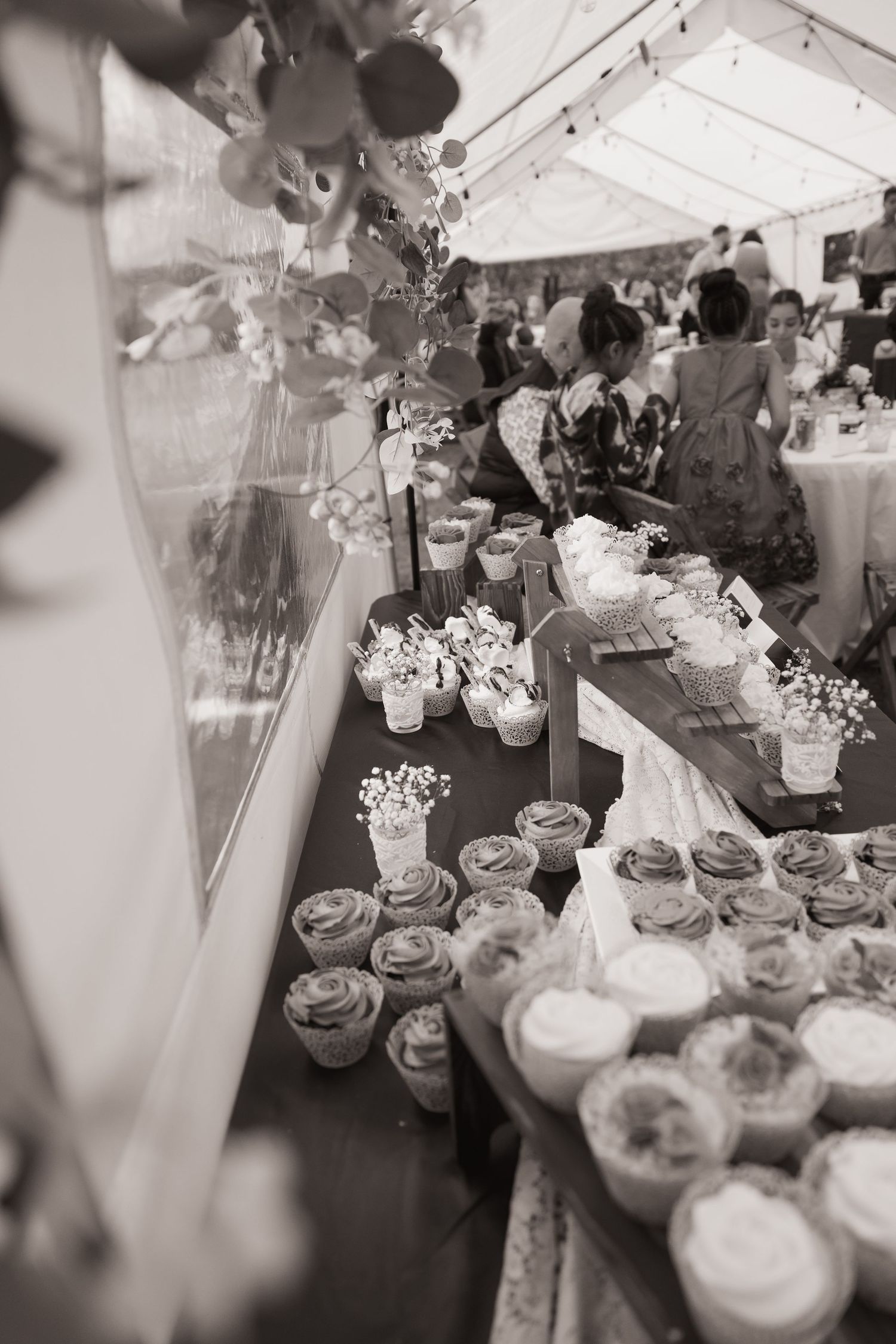 Black and white photo of elegant wedding dessert table with cupcakes and floral decorations under a marquee tent.