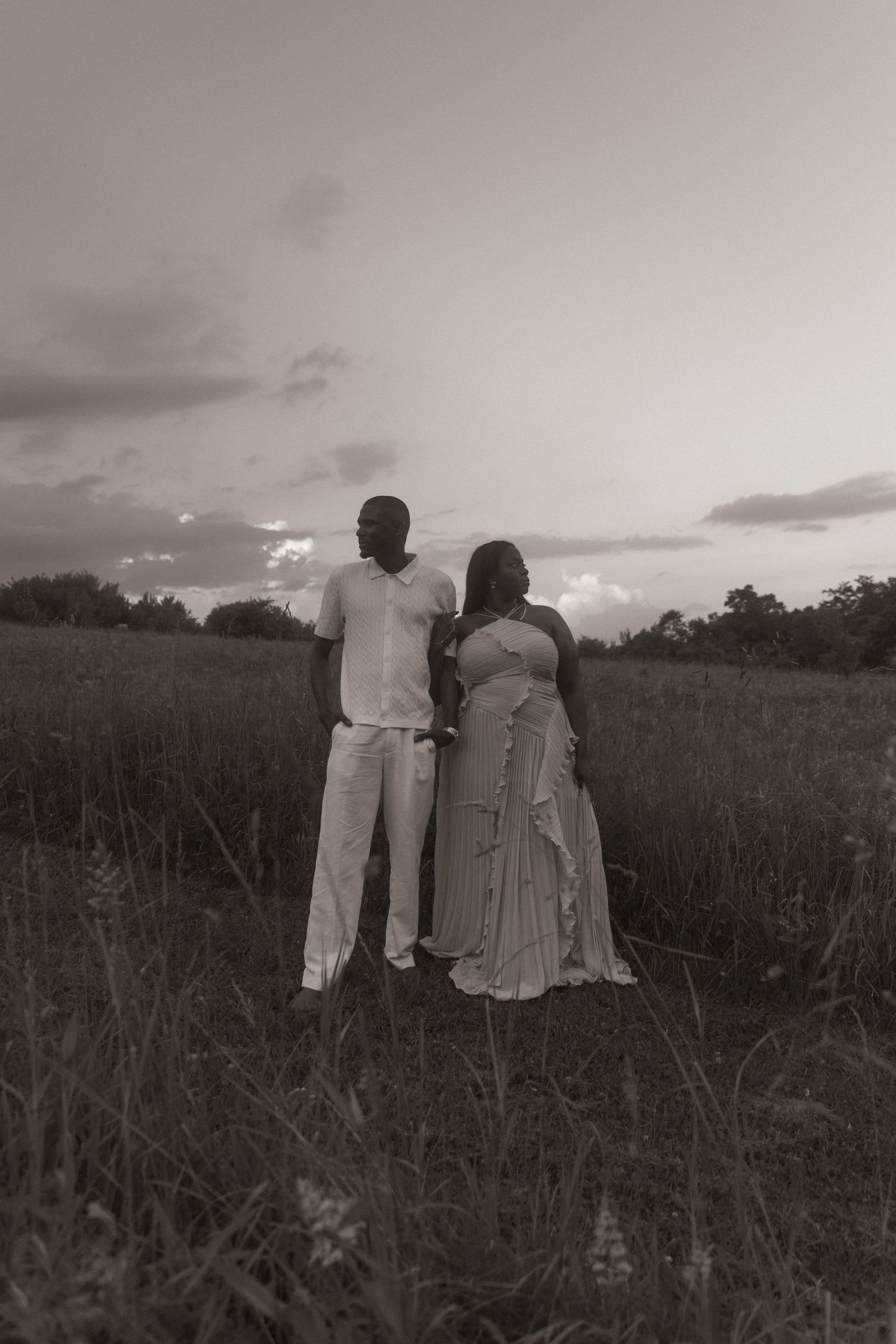 Black and white photo of a couple standing together in a field at sunset, facing away from the camera.