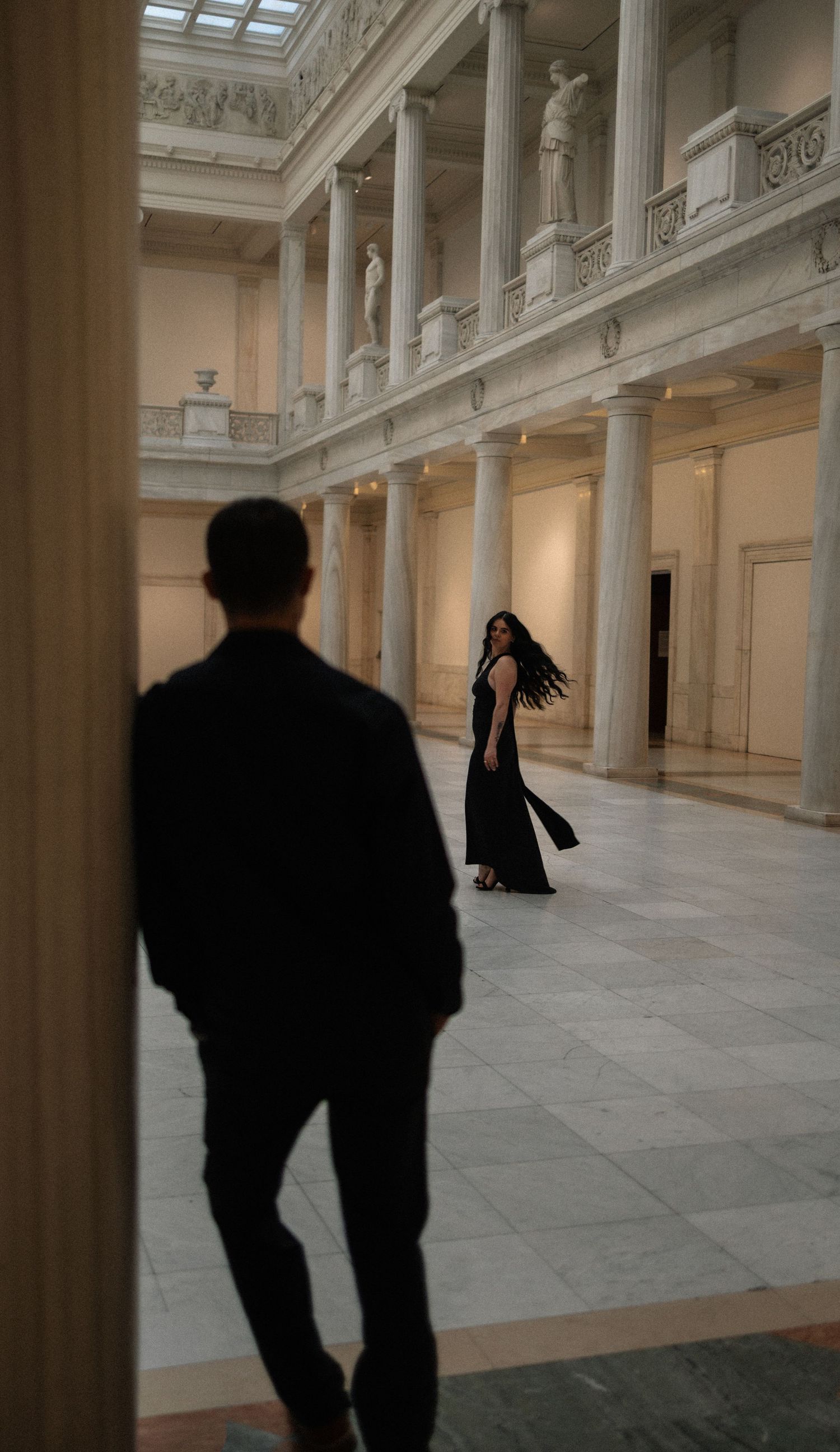 A man in a black suit watches a woman in a black dress twirl in an elegant neoclassical hall with tall columns.