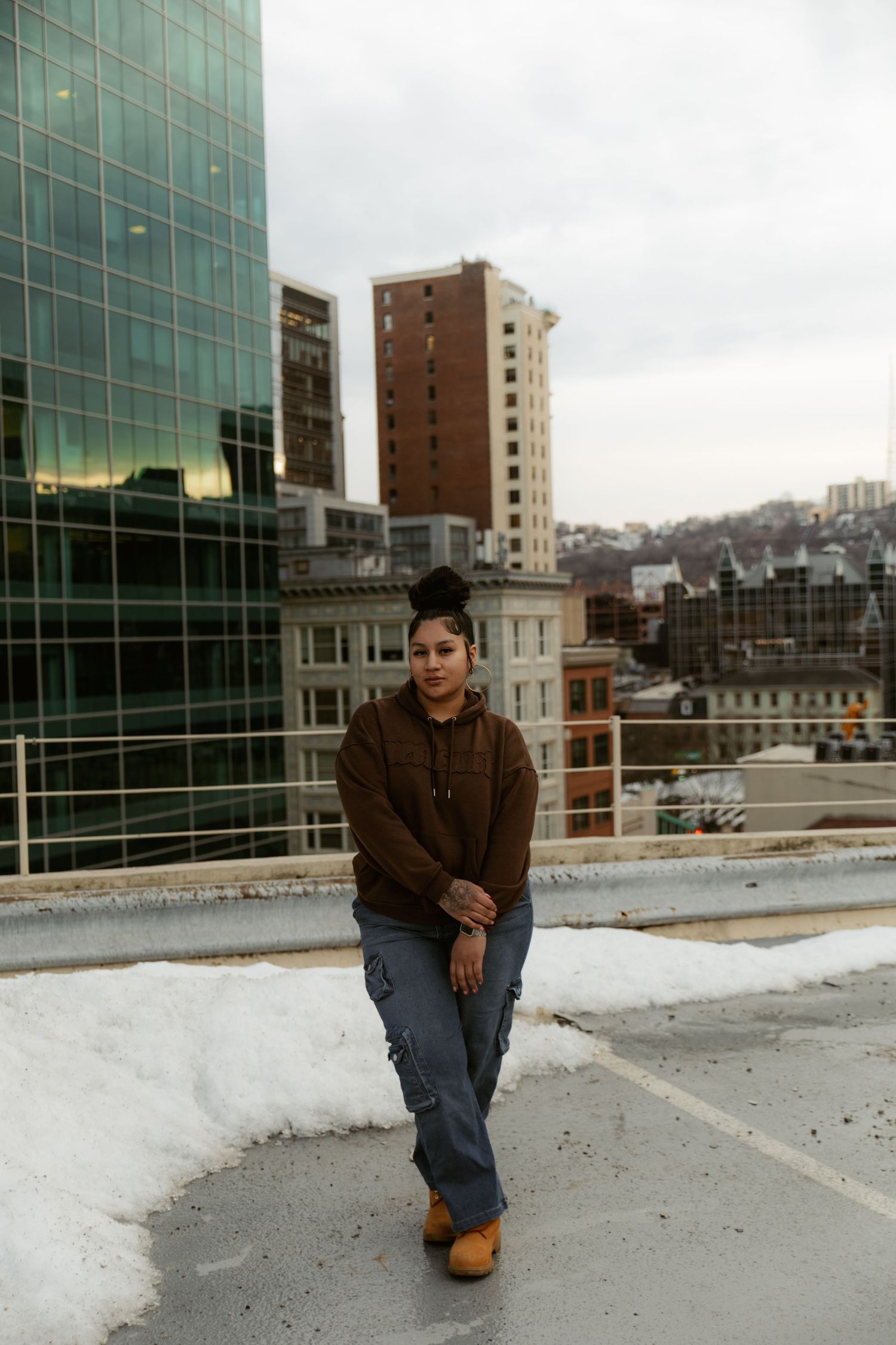 Young woman in brown hoodie and cargo pants poses on urban rooftop with city skyline backdrop in winter.