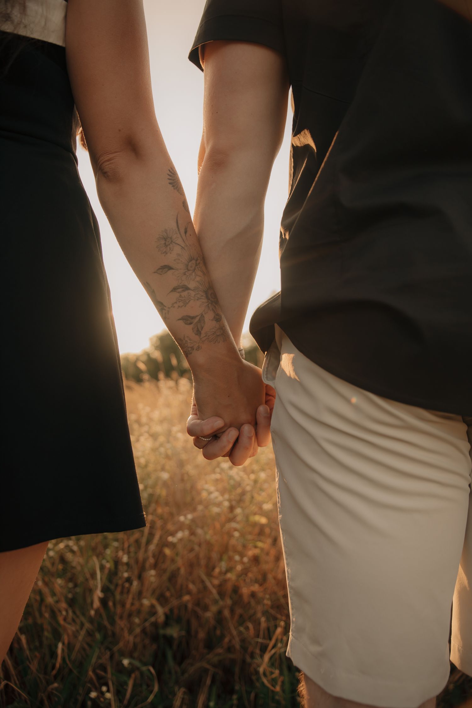 Close-up view of couple holding hands at sunset in a golden wheat field showing tattoos and casual attire.