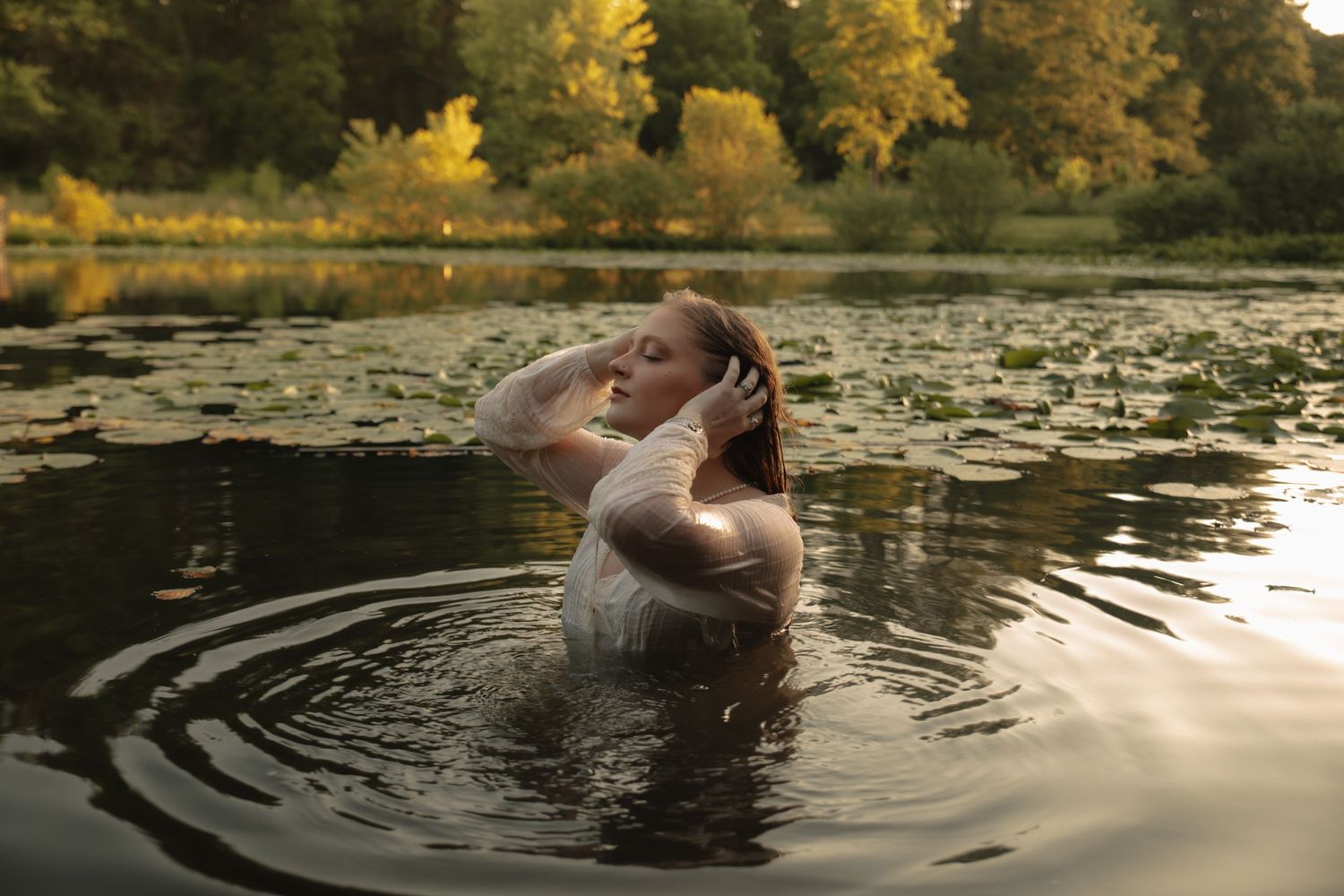 A person emerges from dark water in a lake surrounded by autumn trees, creating concentric ripples on the surface.
