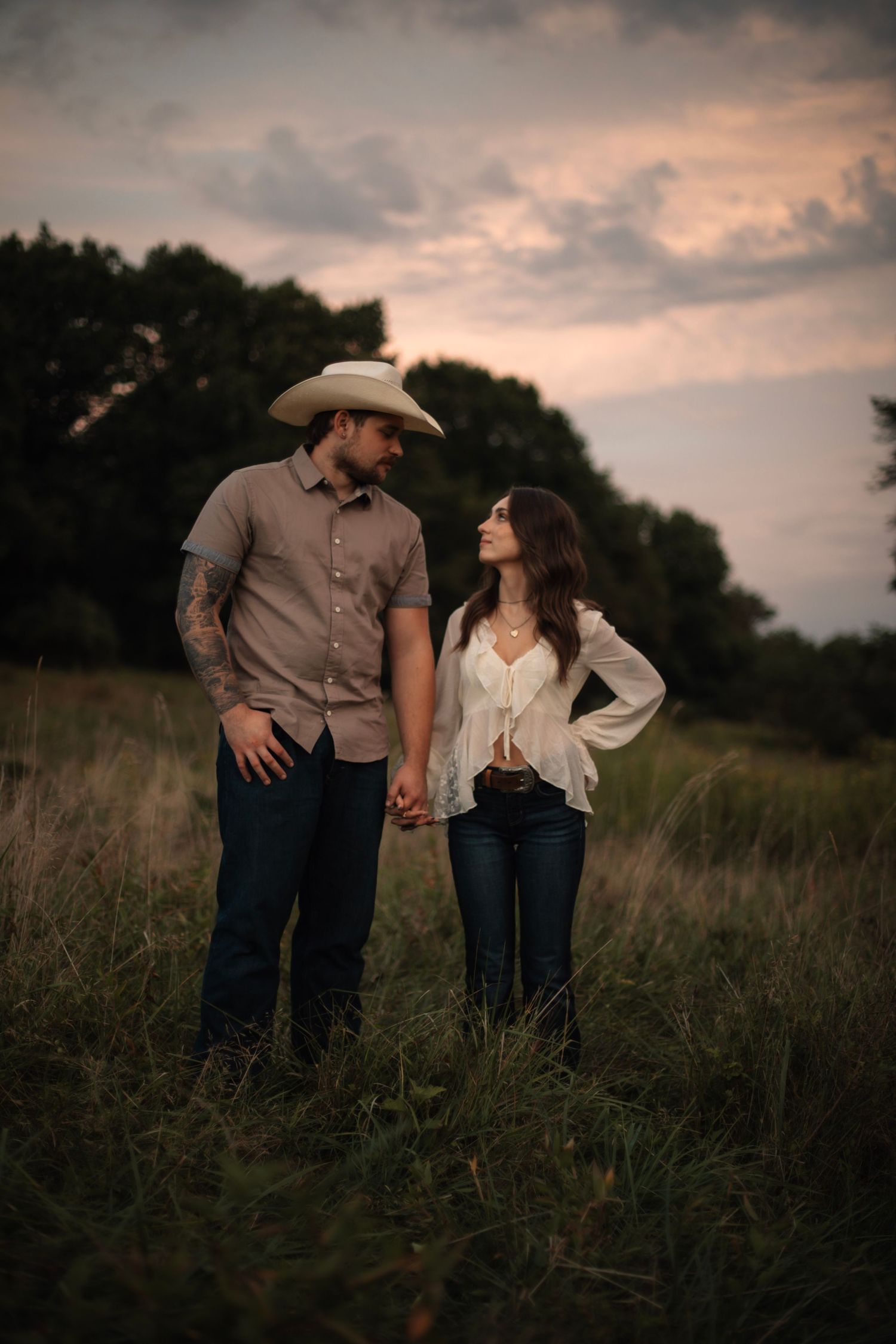 A couple in western wear walks hand in hand through a rustic field during golden hour.