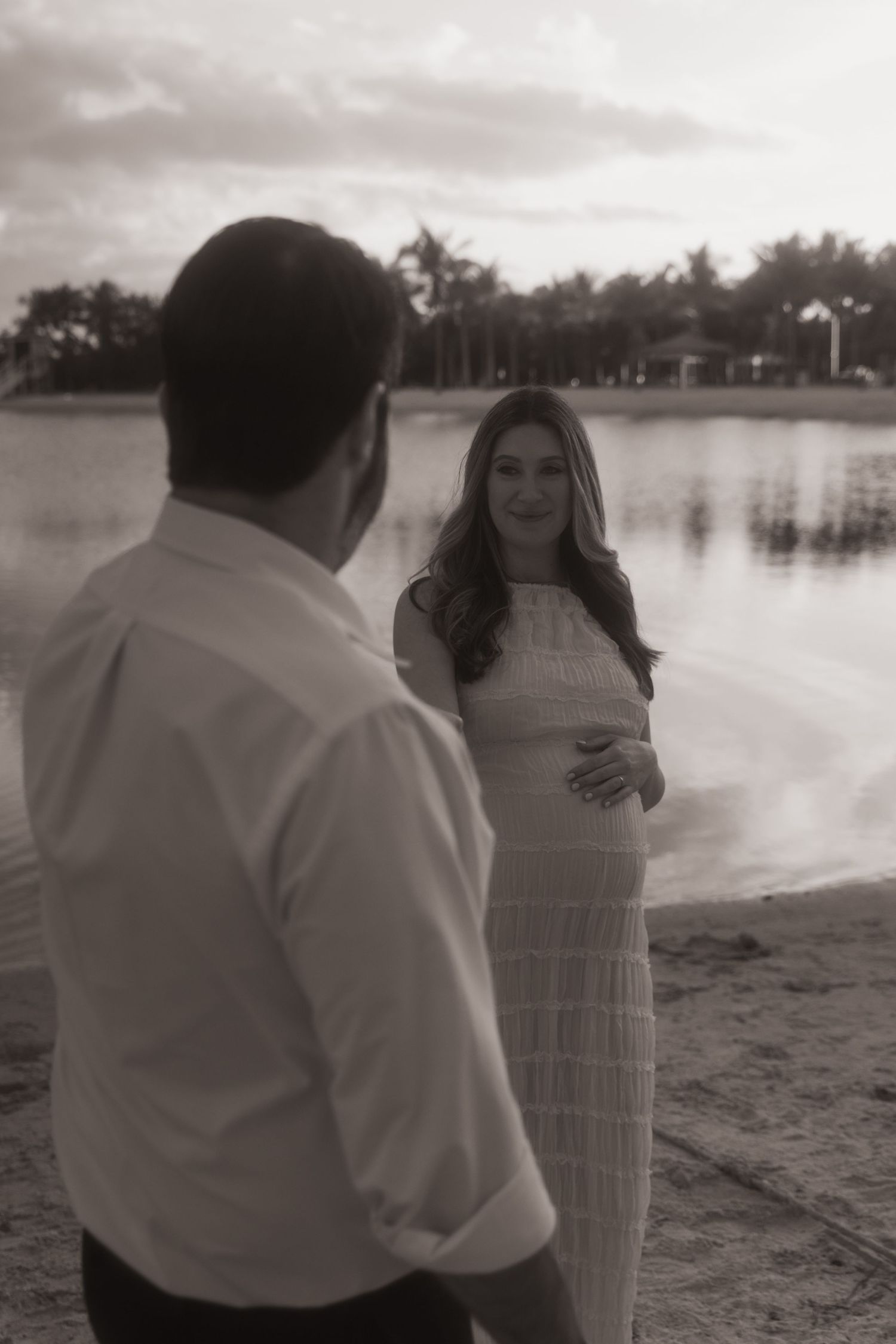 A couple embraces on a beach at sunset with a lake and trees in the background, captured in black and white.
