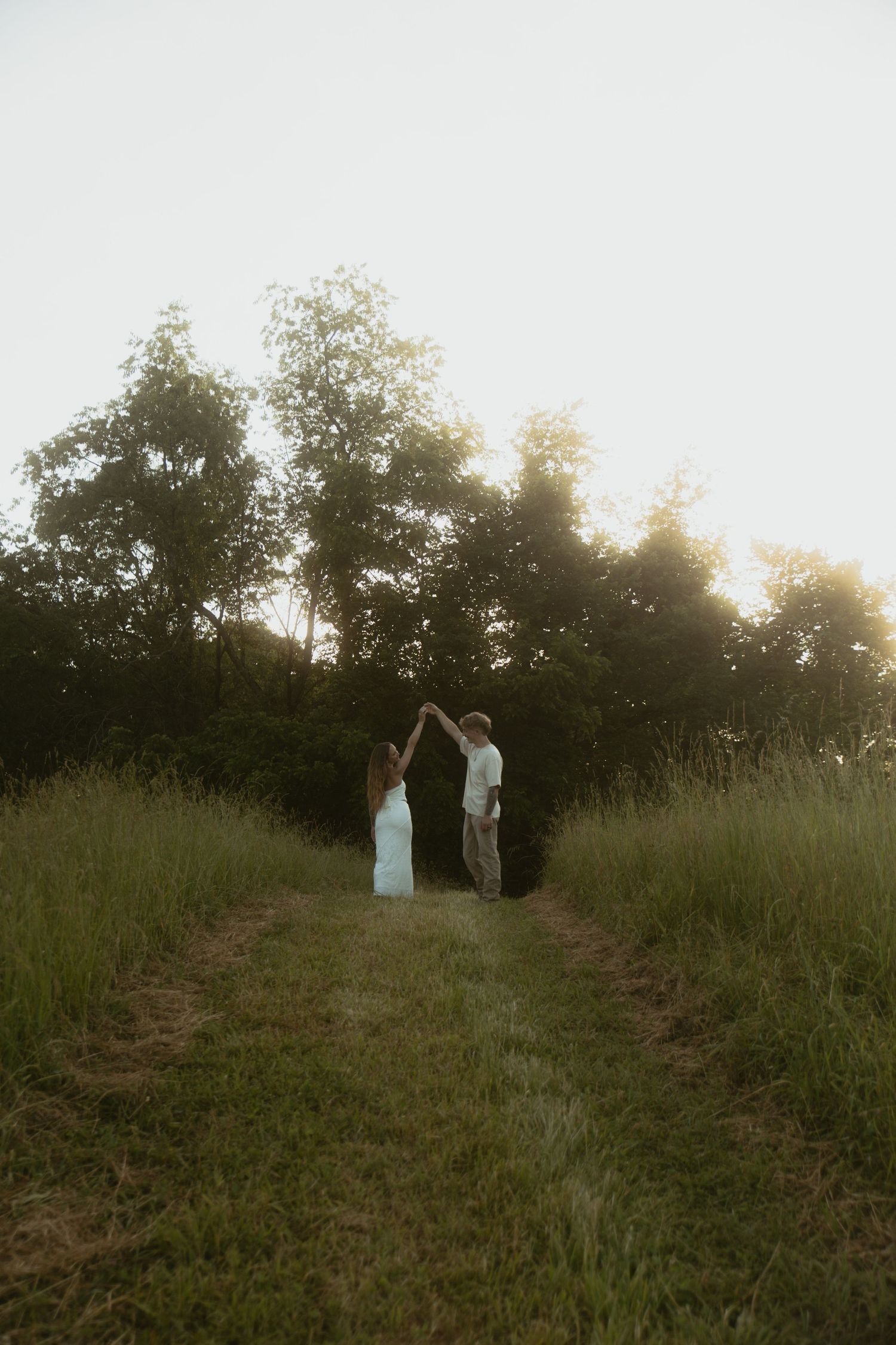 Couple dancing together on a grassy path at golden hour with lush green trees in the background.