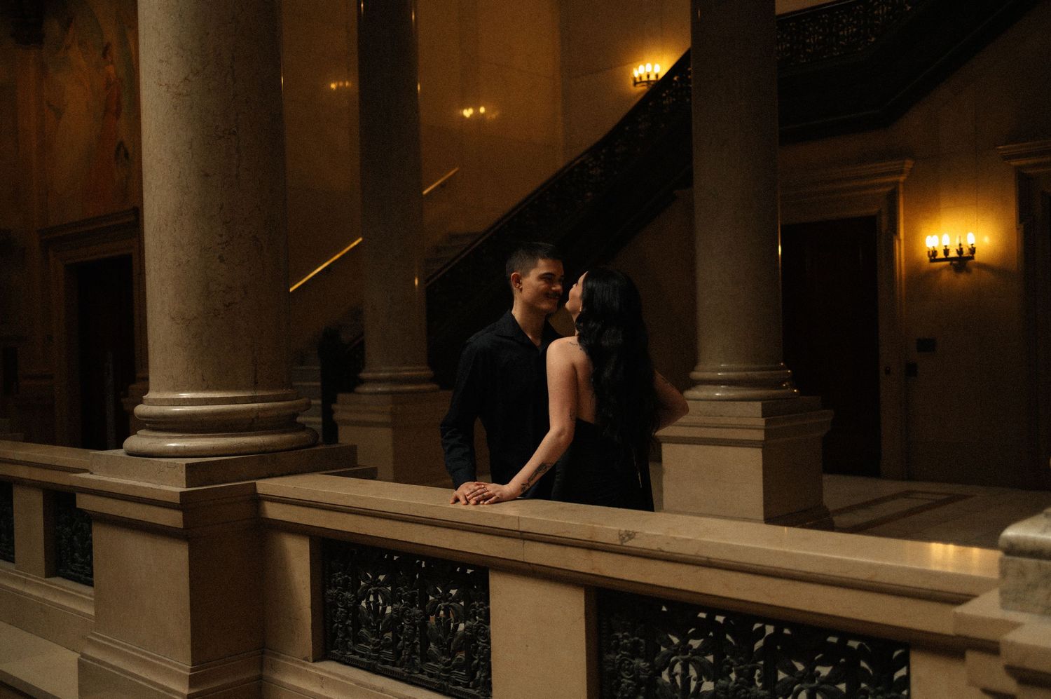 Couple sharing a romantic kiss on an elegant balcony with grand columns and warm lighting in a classic building.
