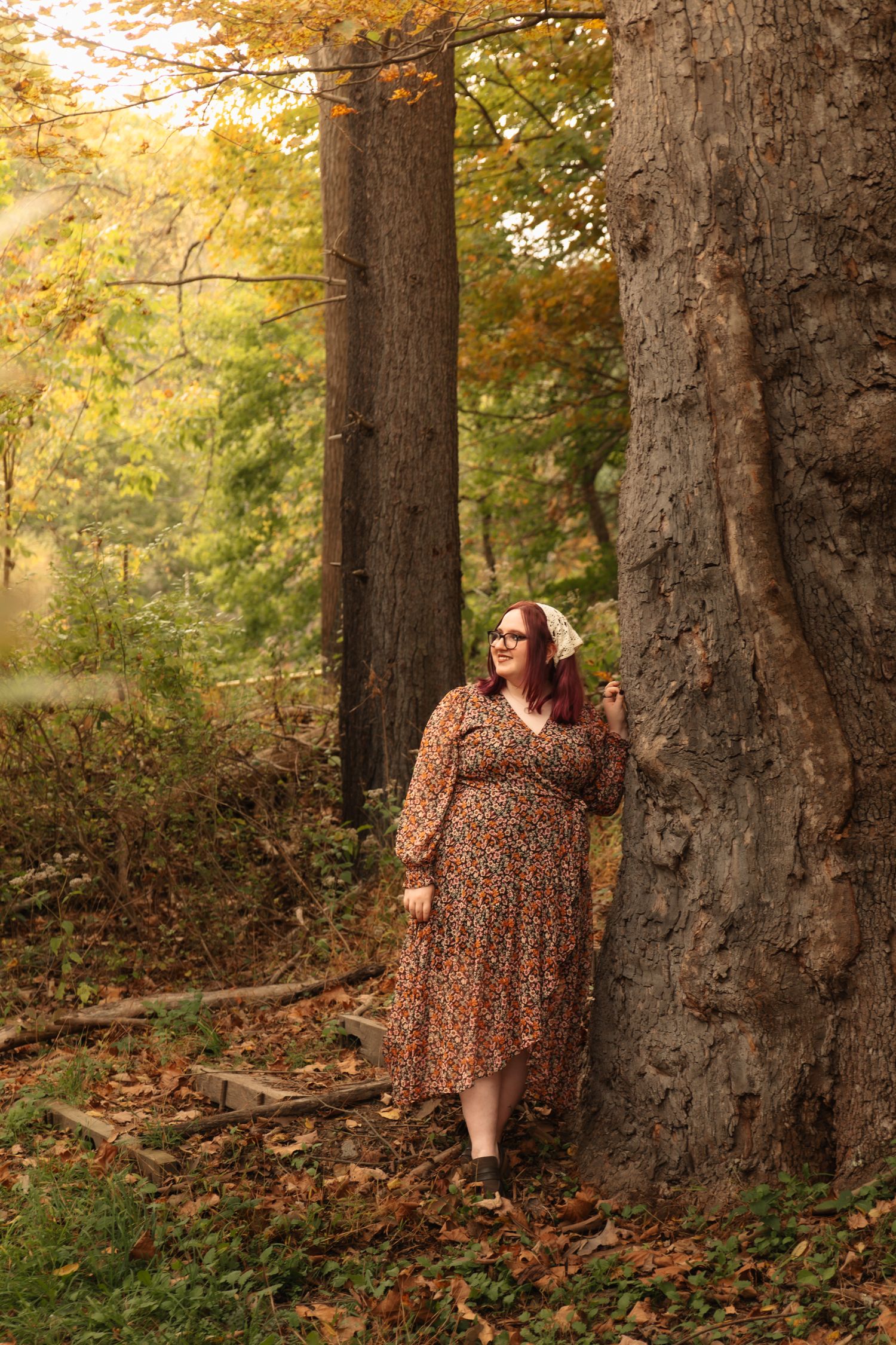 A person in a floral dress sits against large tree trunks in a sunlit autumn forest setting.