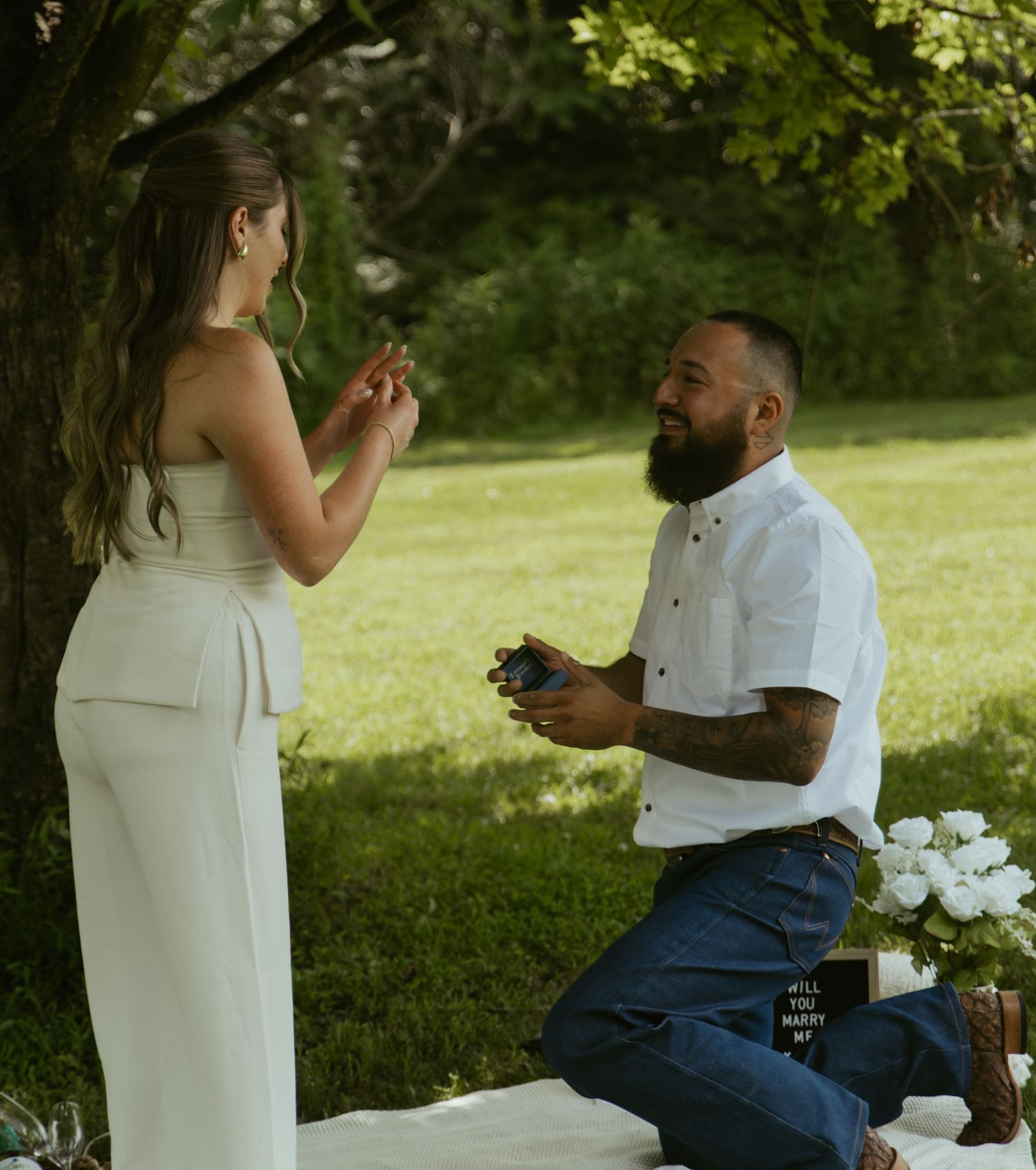 Man kneeling on one knee proposing to woman in white outfit outdoors in green park setting with flowers nearby.