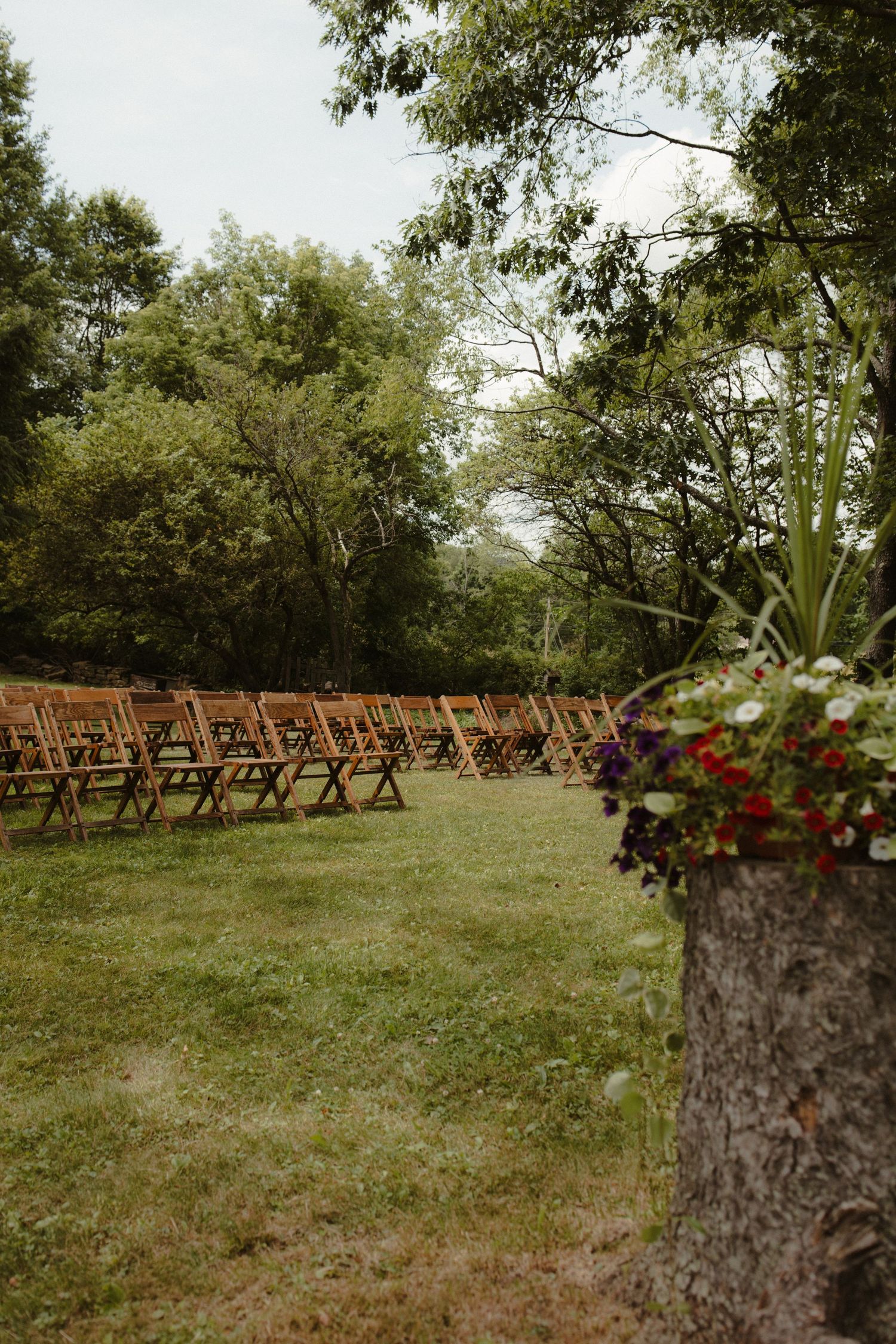 Wooden folding chairs arranged in rows on green grass for an outdoor wedding ceremony, surrounded by lush trees and flower decorations.
