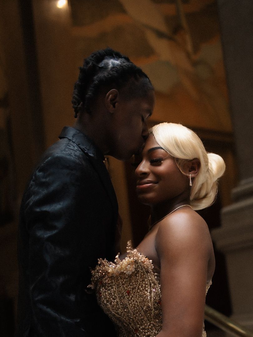 Couple in elegant formal attire share a romantic moment, man in black jacket kisses woman in gold beaded gown.