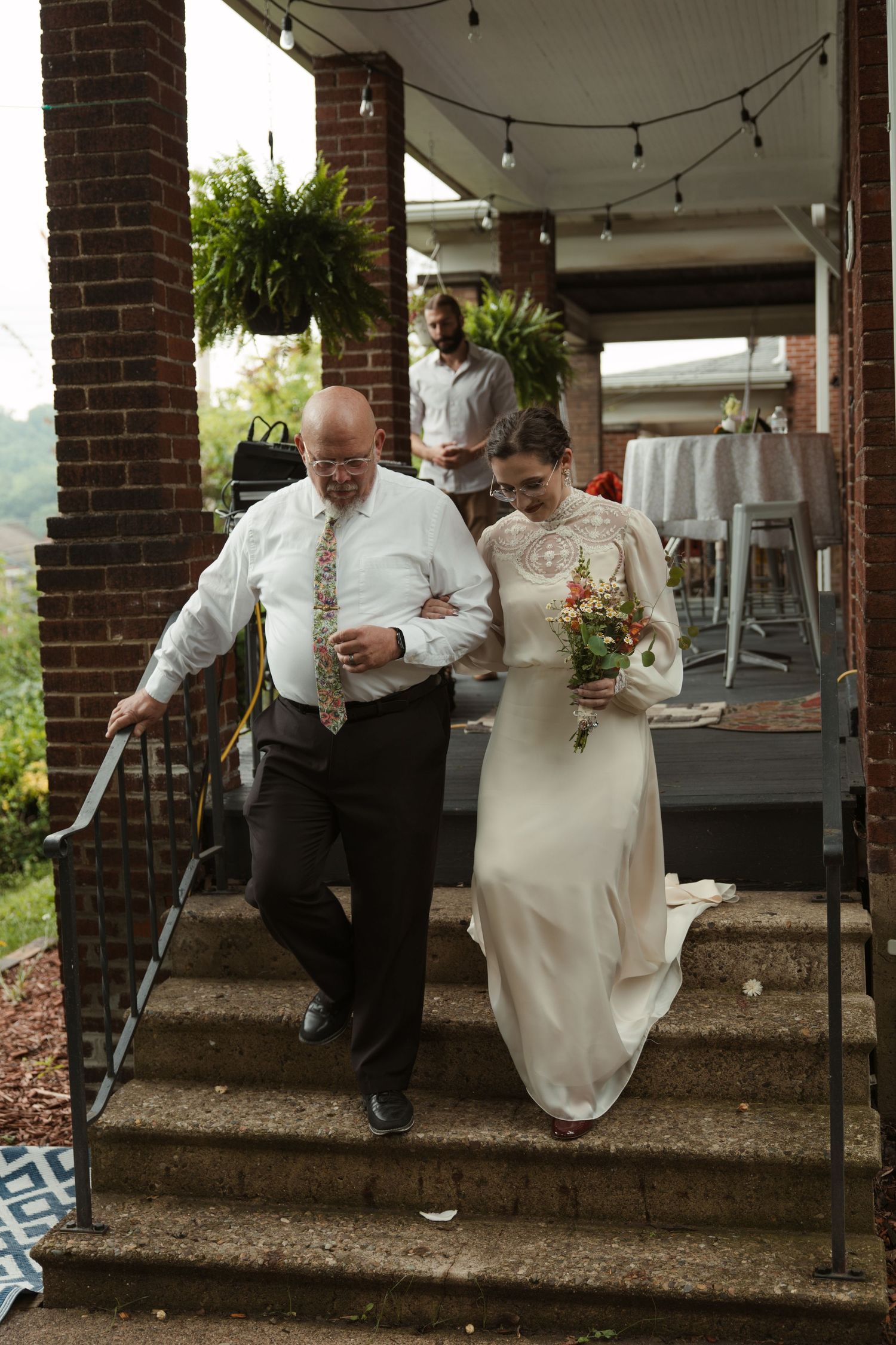 Bride in white gown holding bouquet walks down steps with older man using cane at outdoor wedding venue.
