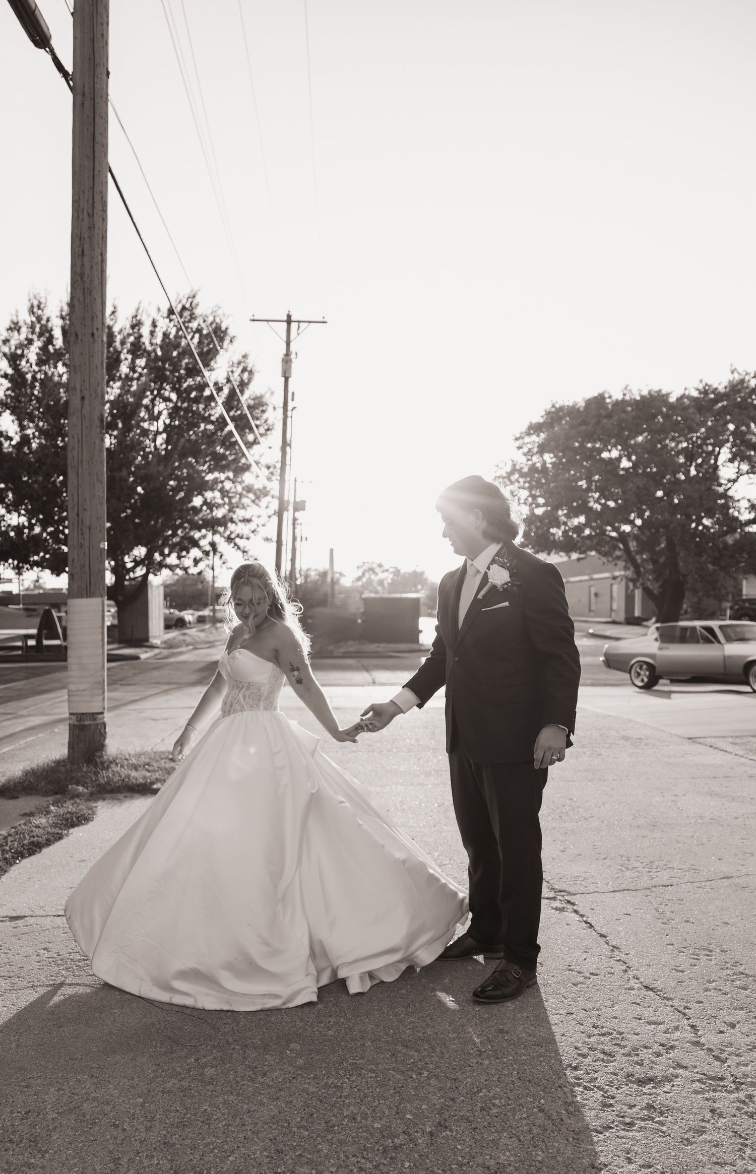 Black and white photo of bride and groom holding hands on a sunlit street, wedding dress flowing gracefully.