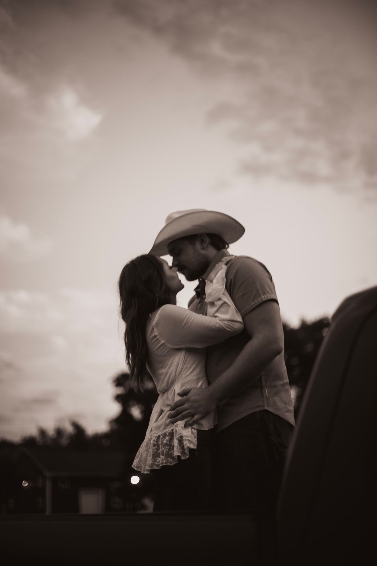 Romantic sepia-toned photo of couple in cowboy hat sharing a kiss at dusk against cloudy sky.
