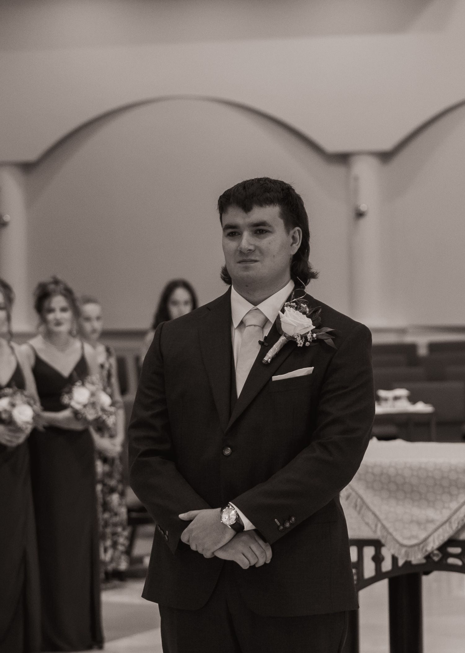 Groom in black suit with boutonniere stands at altar during wedding ceremony, bridesmaids visible in background.