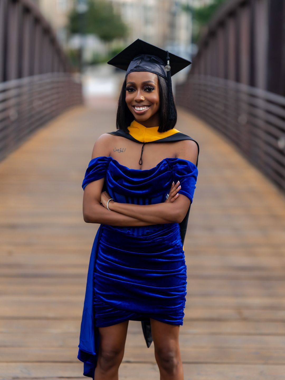 A smiling graduate in a royal blue velvet dress and black cap with gold hood poses on a bridge.
