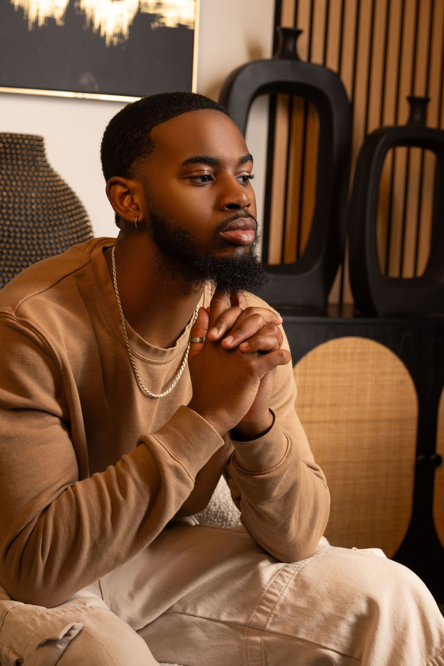 A thoughtful young Black man with a beard sits in a chair, wearing a beige sweatsuit and gold jewelry in a stylish room.