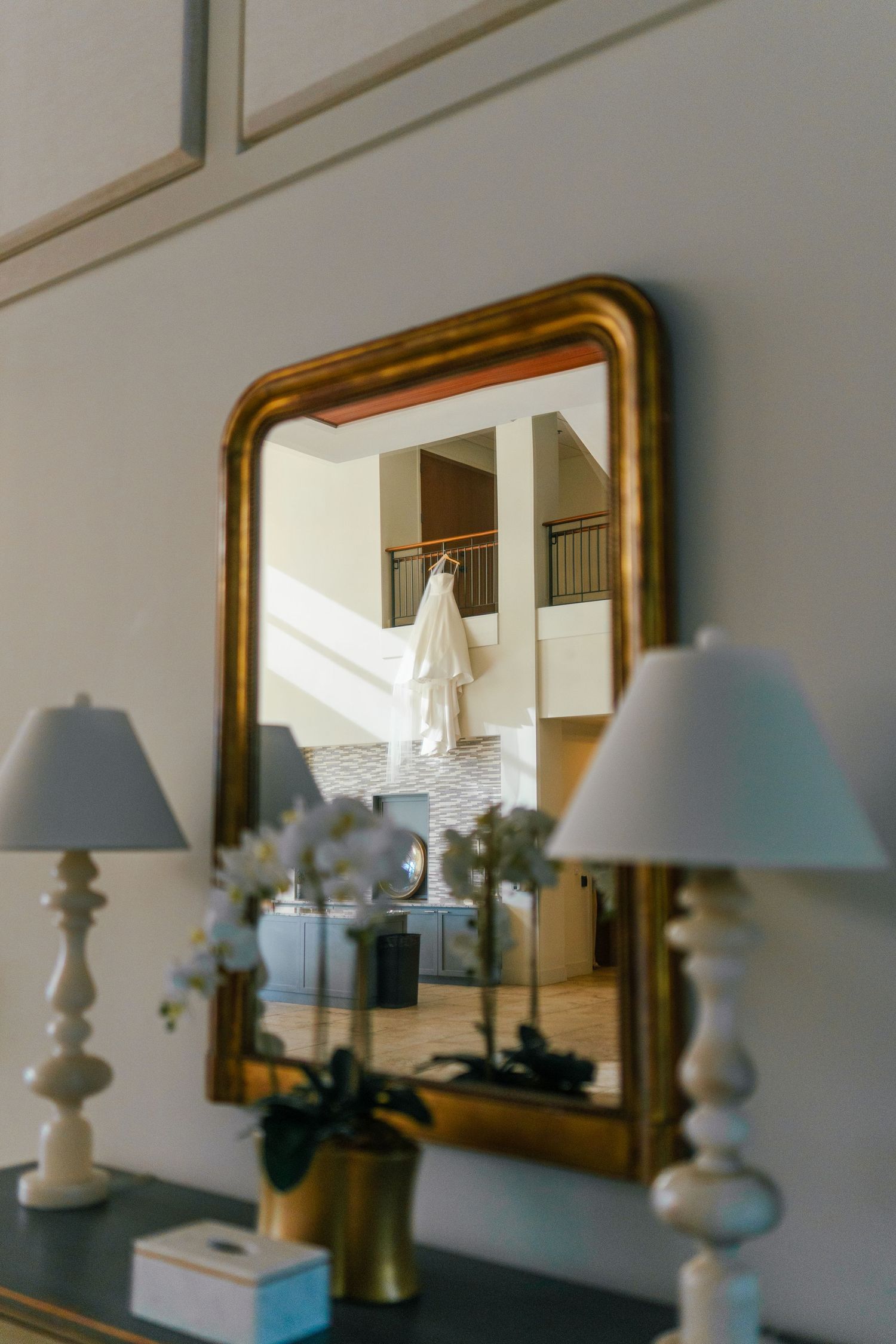 Gold-framed mirror reflecting a wedding dress hanging in a bright foyer, flanked by white lamps and flowers.
