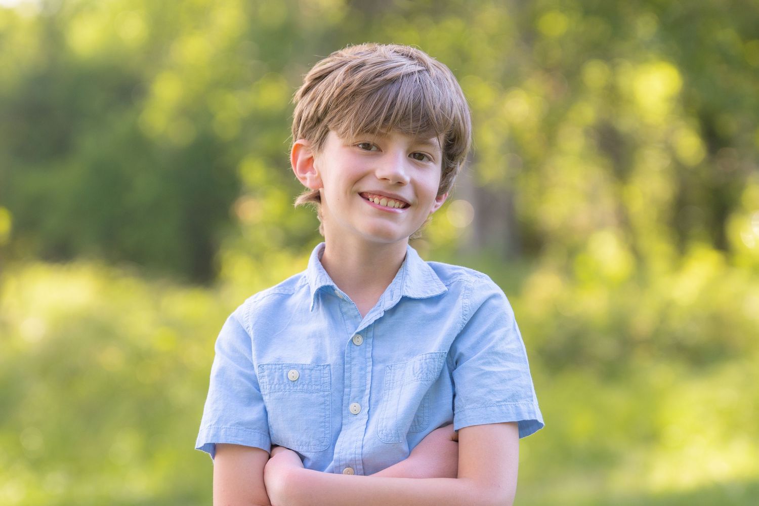 Smiling young boy with crossed arms wearing a blue shirt outdoors in a sunny green park.